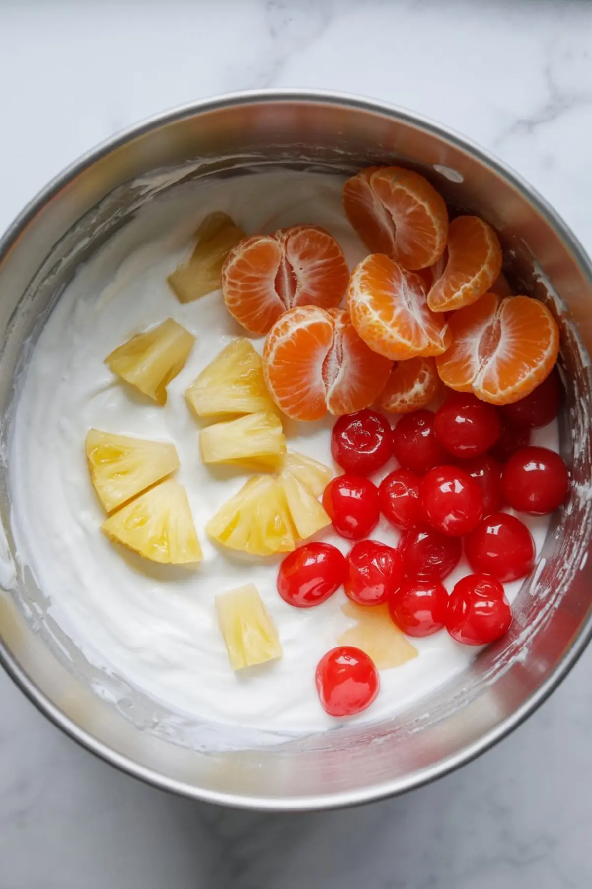 Overhead view of pineapple chunks, mandarin oranges, and maraschino cherries placed over a base of whipped cream in a large metal mixing bowl for ambrosia salad preparation.
