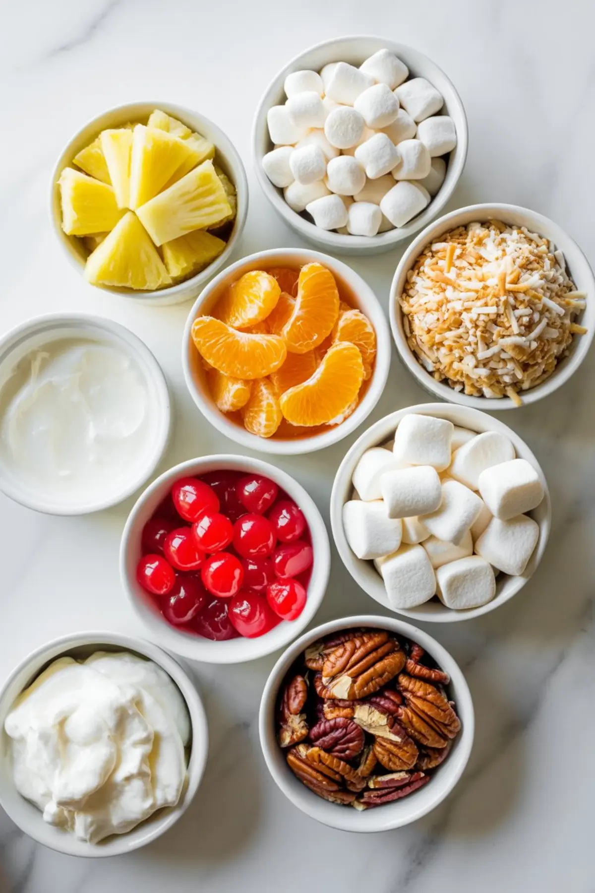 Top-down view of small white bowls filled with ingredients for ambrosia salad, including pineapple chunks, mini marshmallows, shredded coconut, mandarin orange slices, maraschino cherries, pecans, sour cream, and whipped topping.
