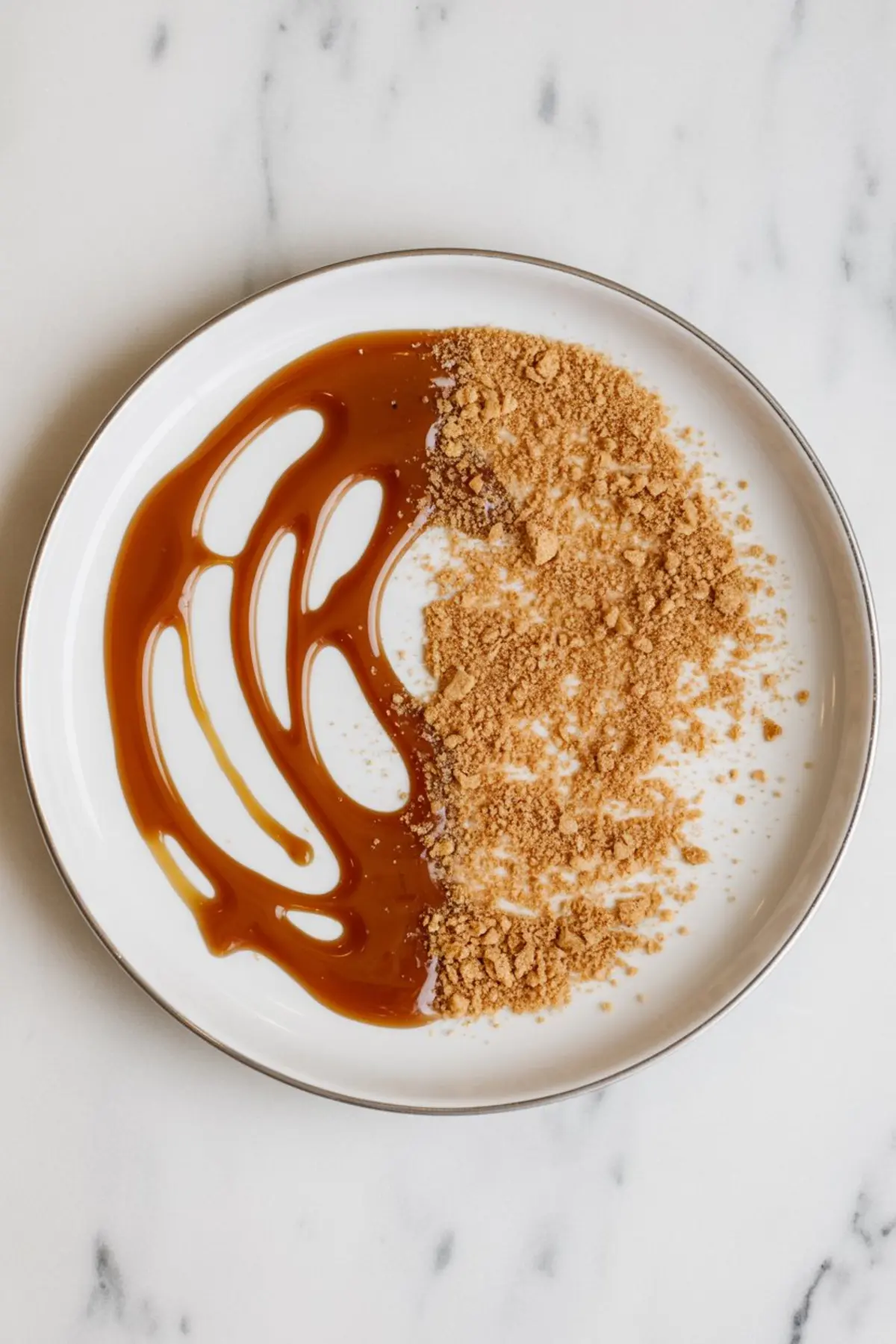 Decorative presentation of caramel sauce and crushed graham cracker crumbs on a white plate with a metallic rim, used for rimming glasses in dessert drinks or cocktails.