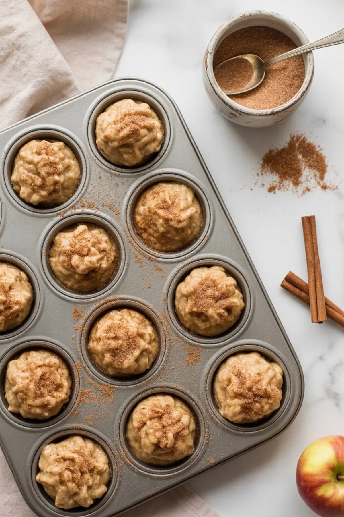 Muffin tin filled with unbaked apple muffin batter, each topped with cinnamon sugar, surrounded by a bowl of cinnamon sugar, whole cinnamon sticks, and a fresh apple.