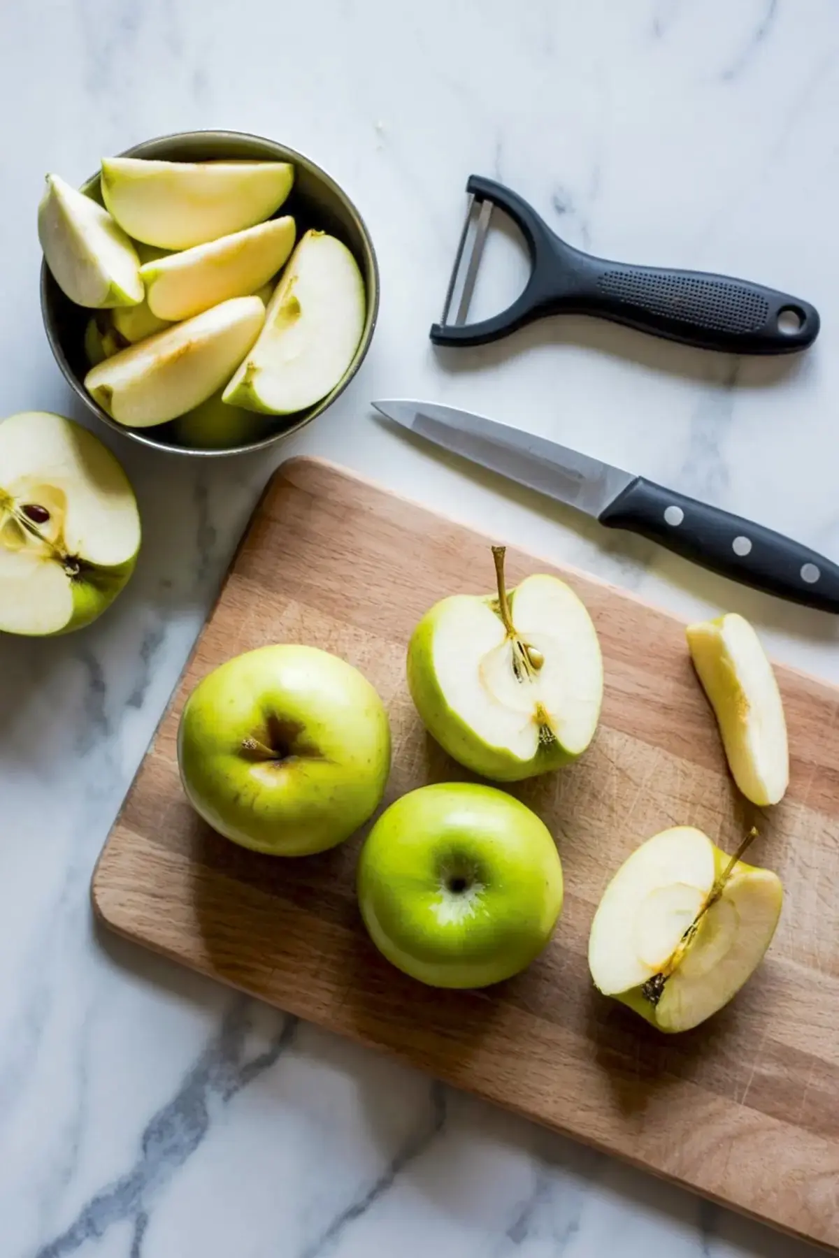 Fresh green apples being sliced on a wooden cutting board with a black-handled knife and peeler, next to a bowl filled with apple wedges.
