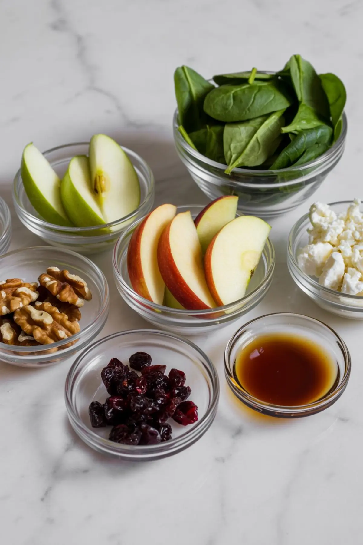 Overhead view of small glass bowls filled with spinach leaves, green and red apple slices, walnuts, dried cranberries, crumbled feta cheese, and balsamic vinaigrette on a white marble surface.