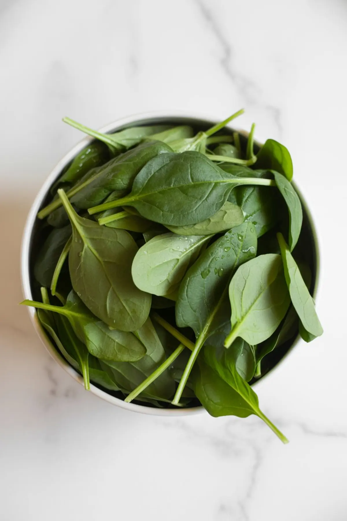 Close-up of fresh baby spinach leaves in a white bowl with water droplets on the surface, set on a marble countertop.