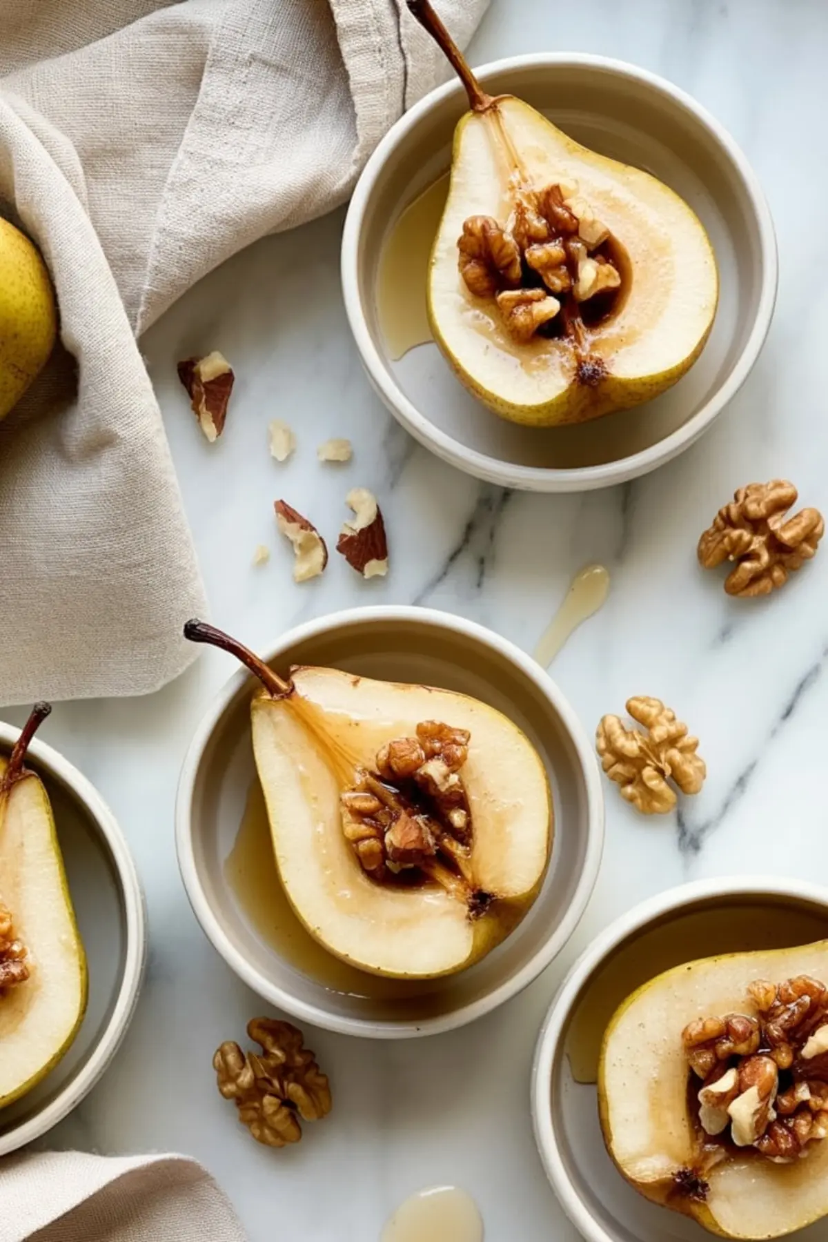 Halved baked pears served in small white bowls, topped with walnuts and drizzled with maple syrup, surrounded by raw walnut pieces on a white marble surface with a beige cloth.