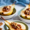 Baked pear halves with maple syrup and walnut filling served in ceramic bowls, with “MAPLE WALNUT BAKED PEARS” text in the upper left over a soft blue background.