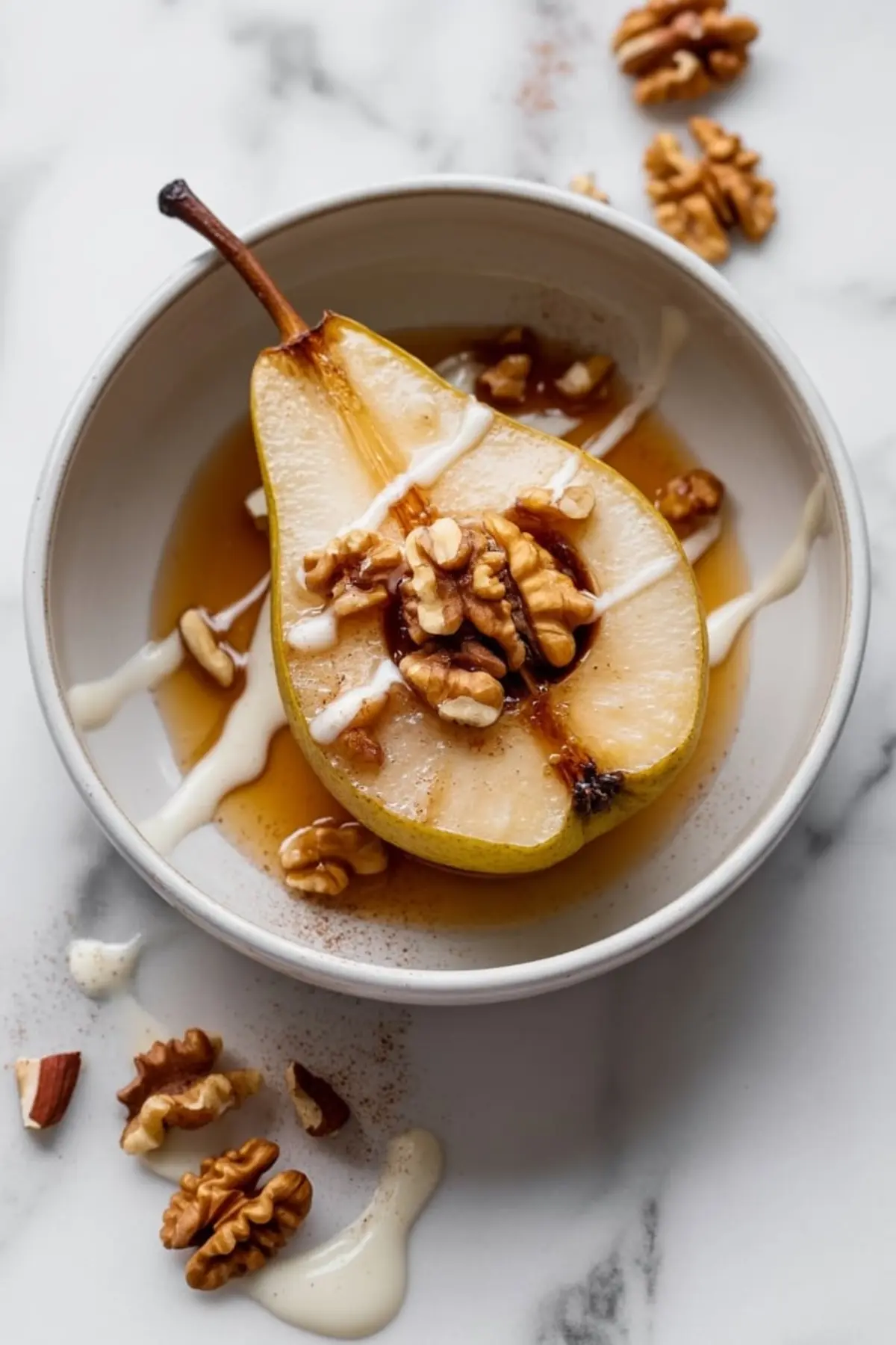 Single baked pear half in a white bowl, topped with walnuts, maple syrup, and white cream drizzle, surrounded by scattered nuts and a light dusting of cinnamon on a marble surface.