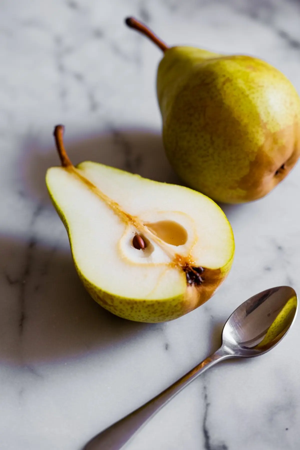 Close-up of a fresh pear sliced in half next to a whole pear and a silver spoon, all placed on a white marble countertop.