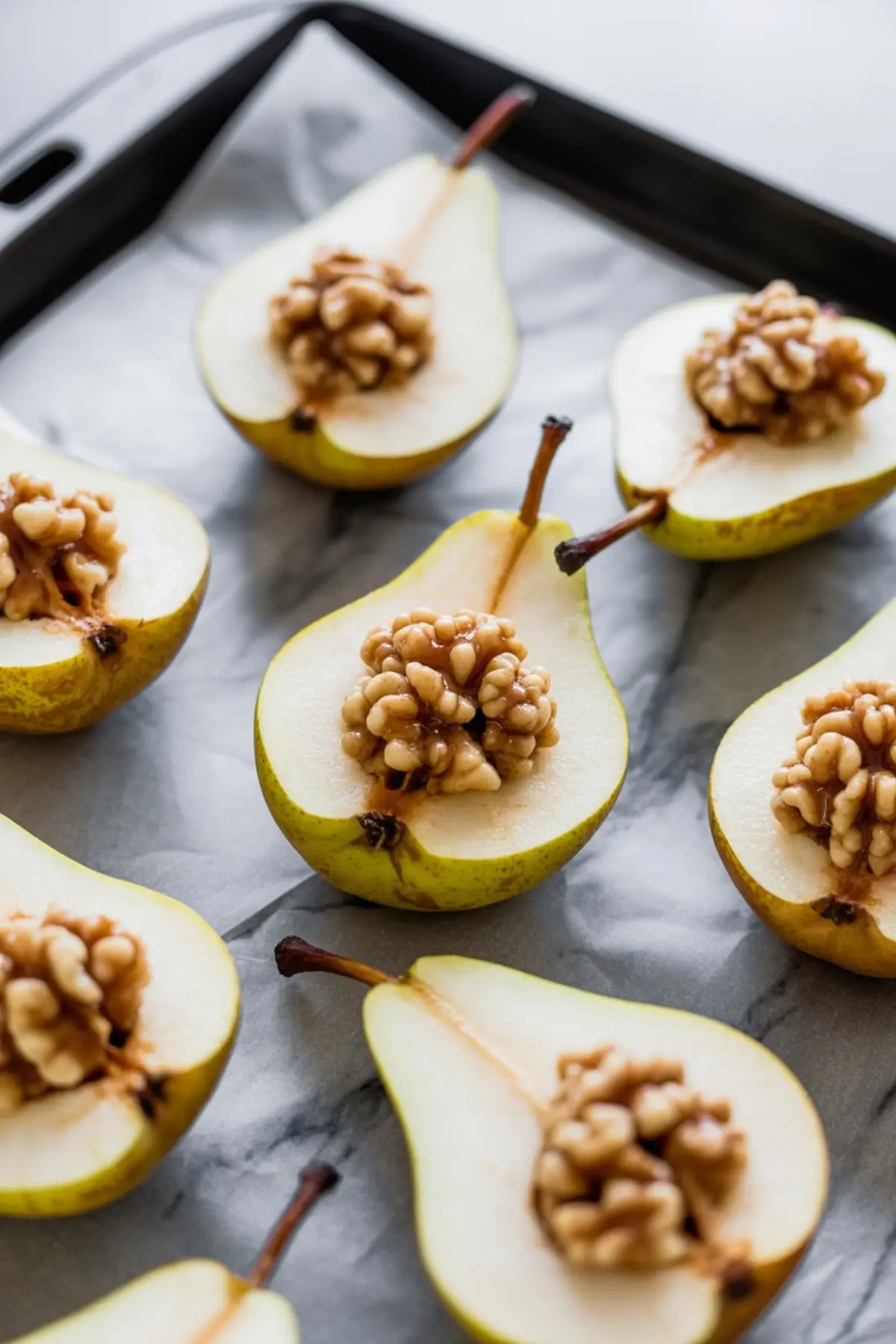 Raw pear halves arranged on a baking sheet, each filled with chopped walnuts and drizzled with maple syrup, ready to be baked.