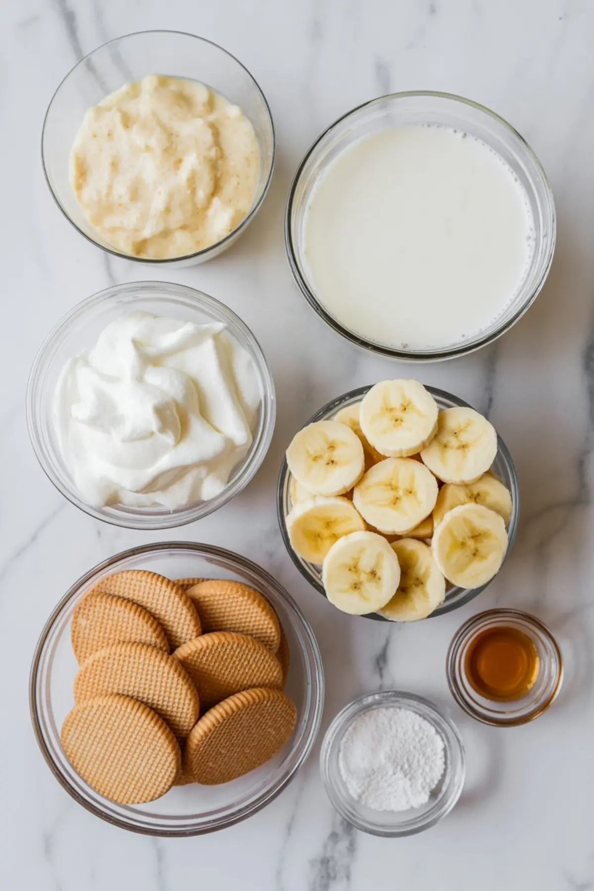 Overhead view of banana pudding ingredients including sliced bananas, vanilla wafers, whipped cream, milk, pudding, sugar, and vanilla extract in glass bowls on a marble surface.