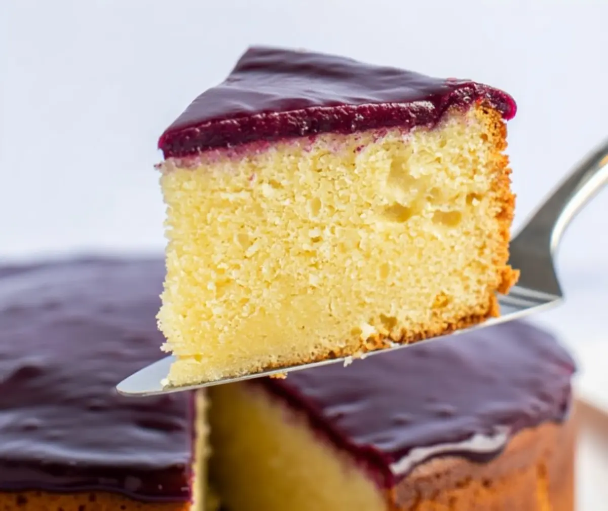 Close-up of a single slice of yellow cake with a moist crumb, topped with a thick, glossy layer of berry glaze, being lifted from the whole cake in the background.
