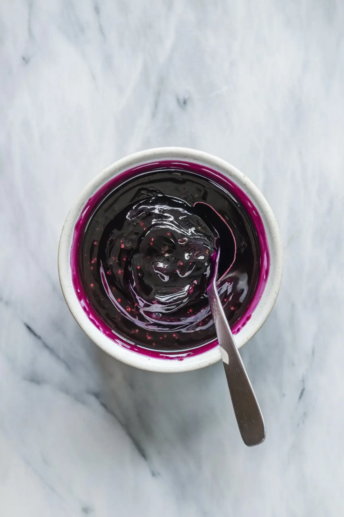 White ceramic bowl filled with glossy berry compote or mixed berry glaze with visible seeds, and a spoon resting inside, placed on a marble surface.