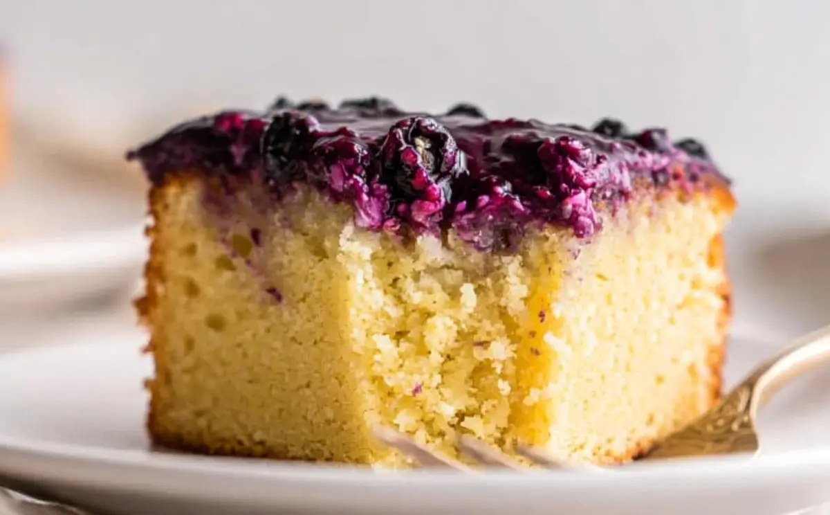 Close-up of a square slice of yellow cake topped with chunky blueberry glaze, served on a white plate with a dessert fork.