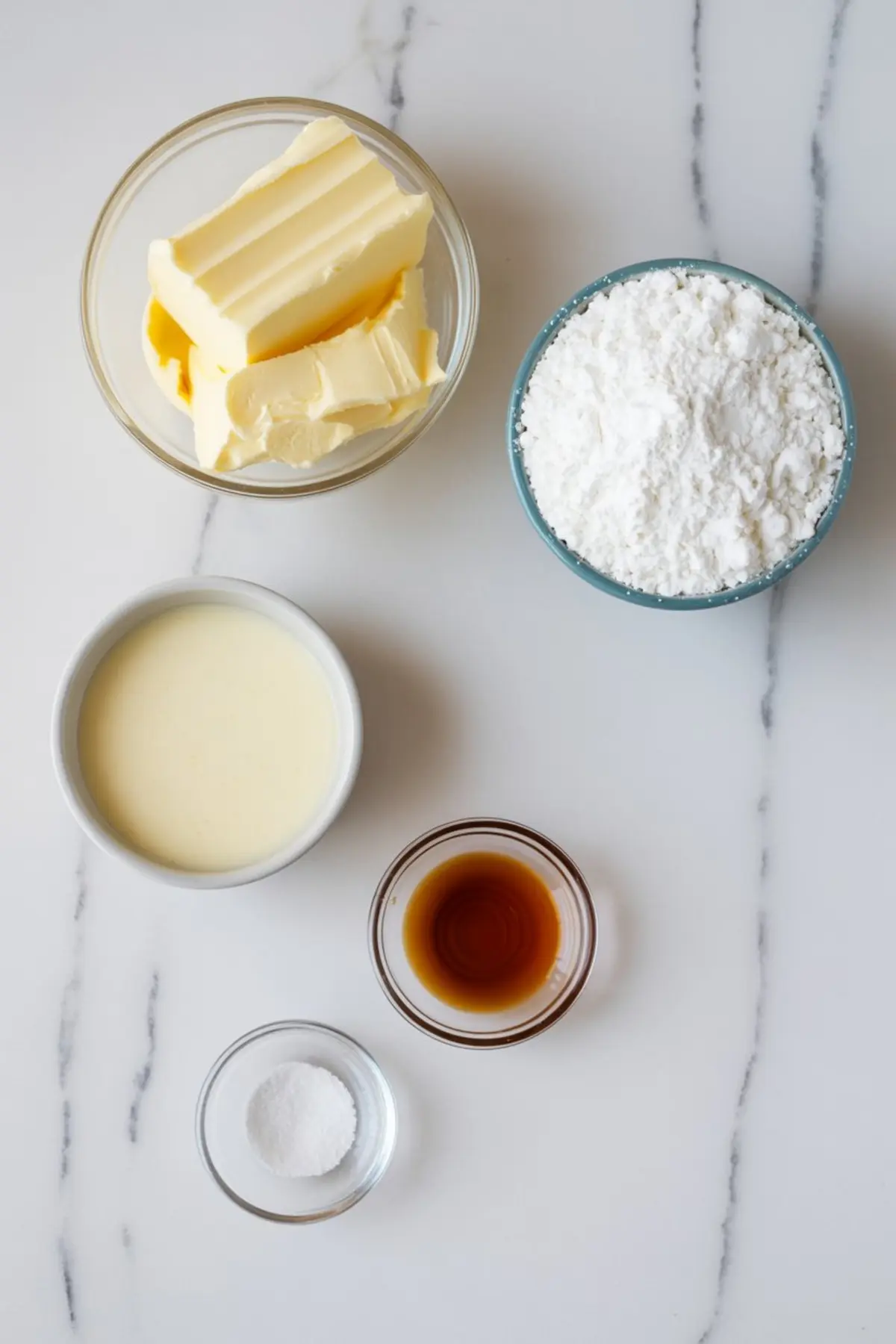 Flat lay of buttercream frosting ingredients including butter, powdered sugar, vanilla extract, milk, and salt in small bowls on a marble surface.