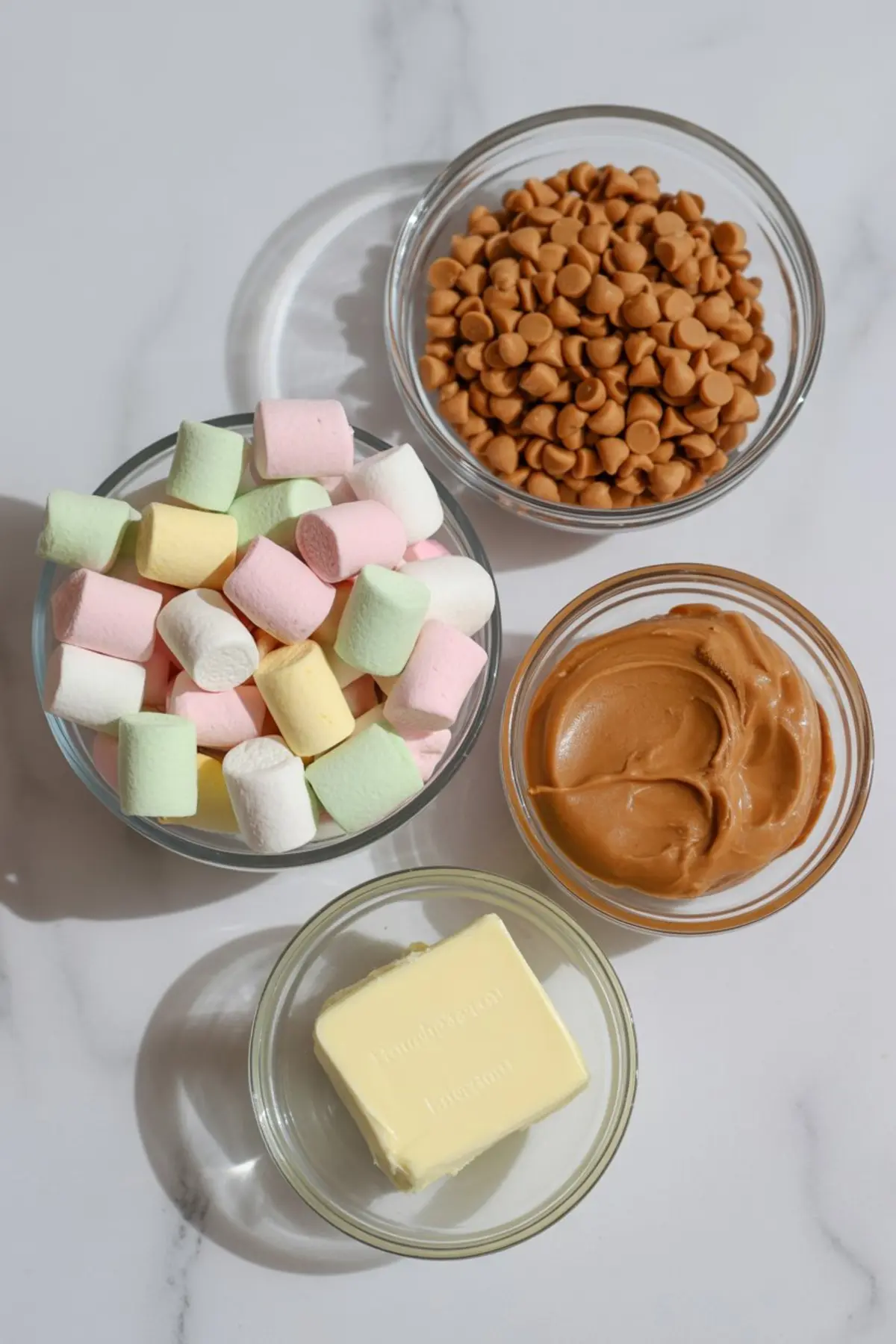 Flat lay of butterscotch marshmallow square ingredients in glass bowls, including pastel mini marshmallows, butterscotch chips, peanut butter, and butter.