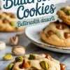 Buttery pistachio butterscotch cookie on marble with melted butterscotch chips and pistachio pieces, surrounded by whole pistachios and a plate of cookies in the background with text overlay “Butterscotch Cookies – Butterscotch desserts.”