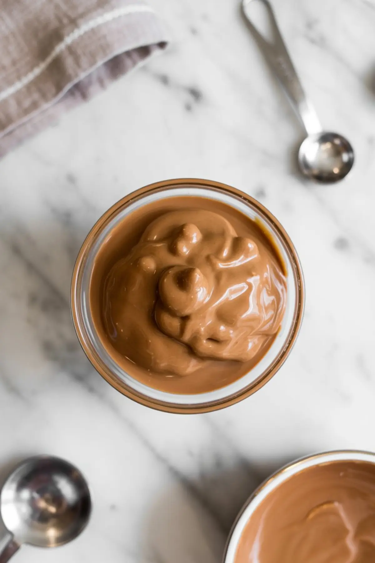 Overhead view of a bowl filled with smooth, creamy melted butterscotch on a marble countertop with measuring spoons nearby.