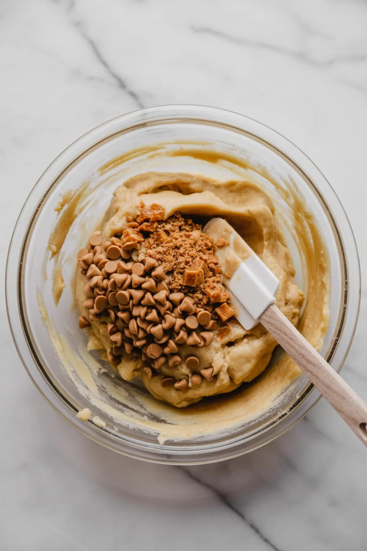Partially mixed cookie dough in a clear bowl with butterscotch chips and chopped toffee pieces sitting on top, ready to be folded in with a rubber spatula.

