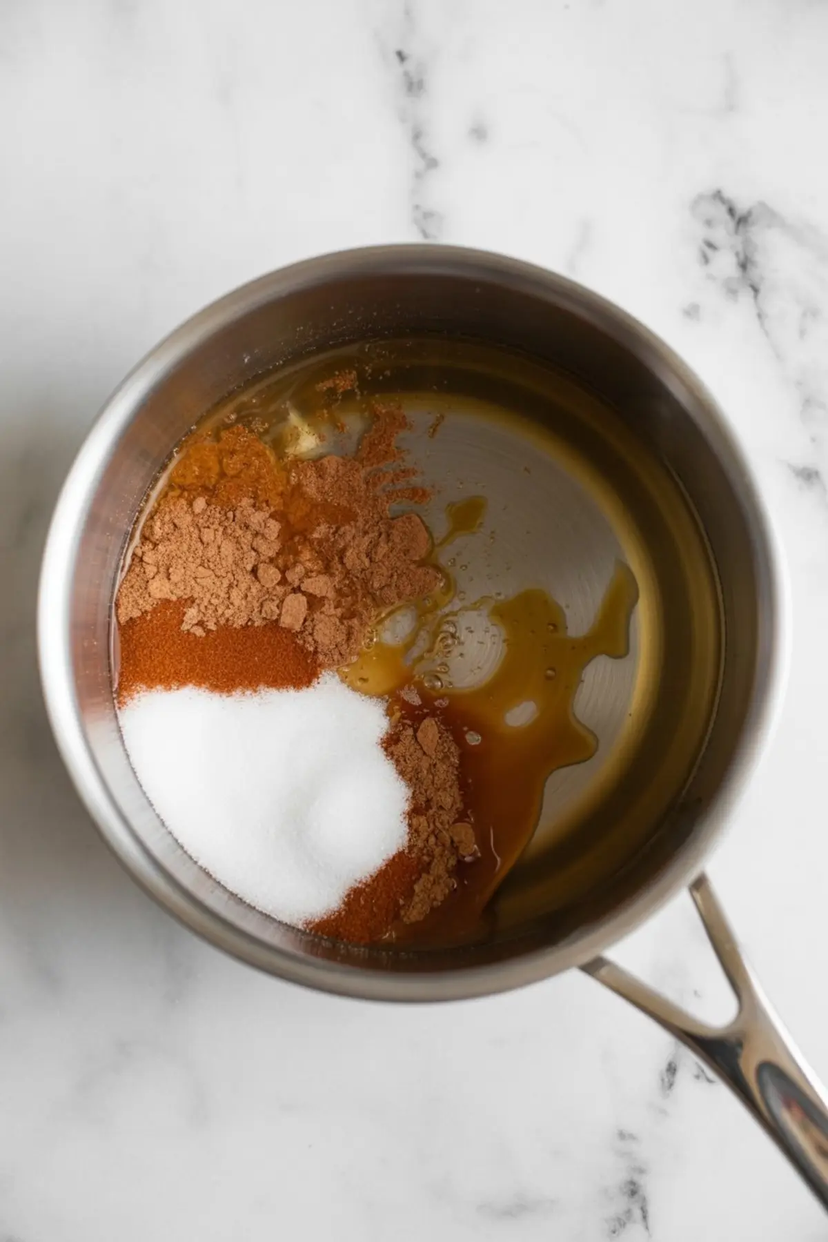 Unmixed ingredients including white sugar, cinnamon, nutmeg, vanilla extract, and oil sitting in a stainless steel saucepan, ready to be stirred for a candied walnut recipe.