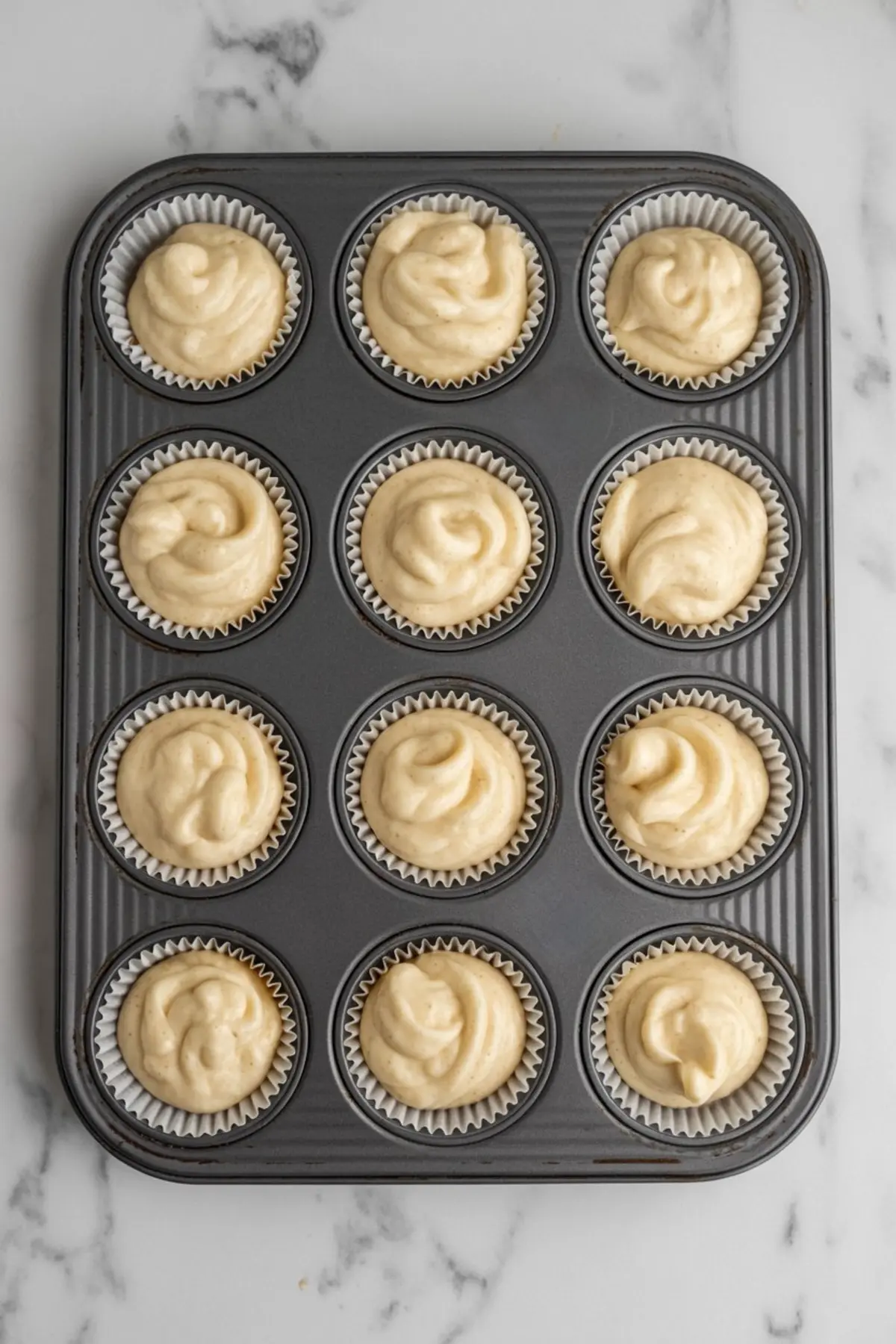 Overhead view of a cupcake tray filled with raw vanilla batter in paper liners, ready to be baked into fluffy cupcakes.