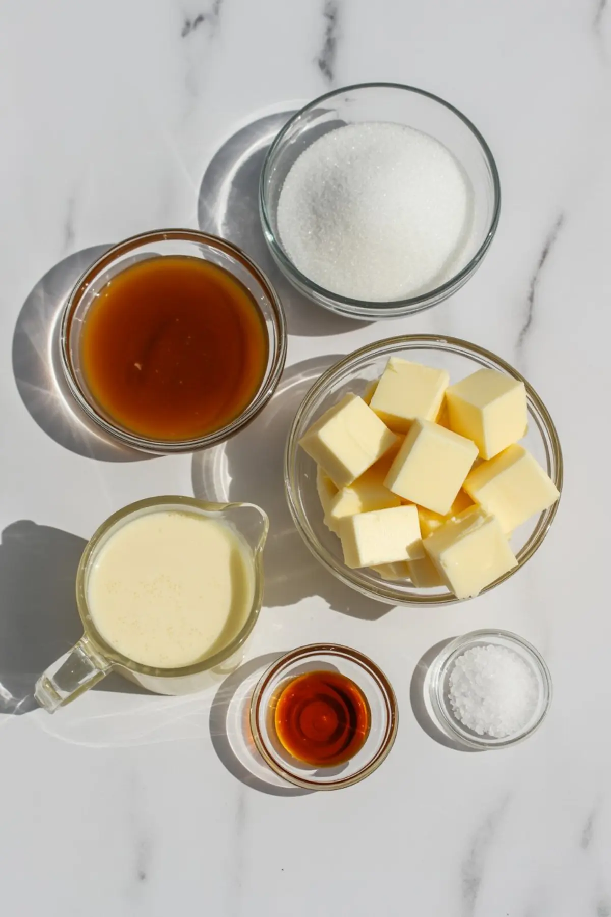 Flat lay of caramel sauce ingredients including butter cubes, sugar, heavy cream, vanilla extract, sea salt, and caramelized sugar in glass bowls on a marble surface.