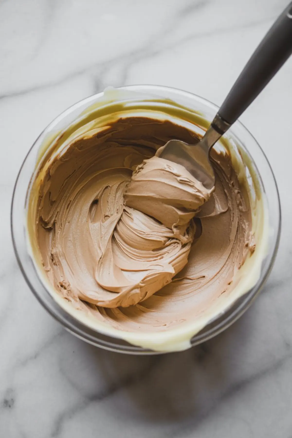 Smooth coffee frosting in a mixing bowl, whipped to a creamy, fluffy consistency with a spatula dipped into the swirl of mocha-colored frosting.