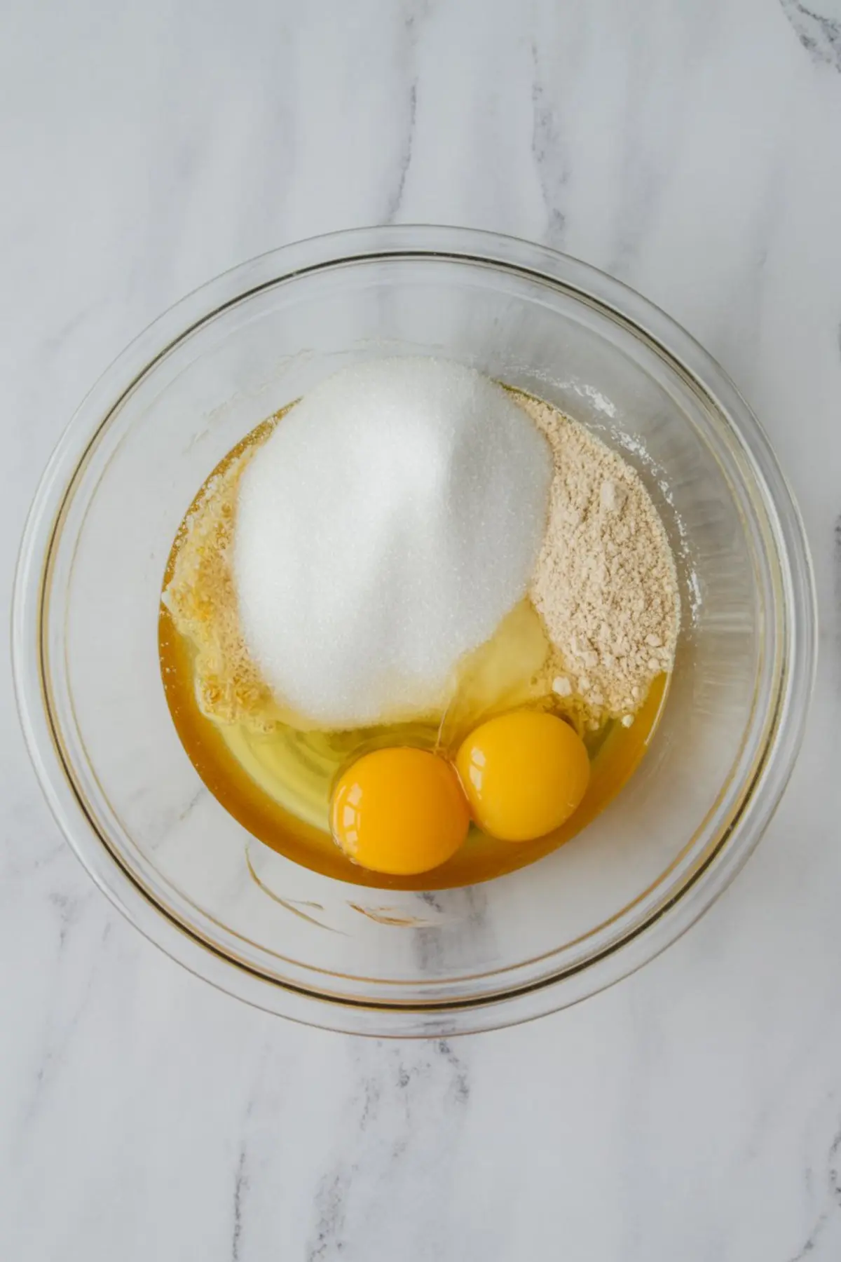 Overhead view of raw cake ingredients in a clear mixing bowl, featuring two egg yolks, granulated sugar, flour, and vanilla on a marble surface, ready for blending into cake batter.
