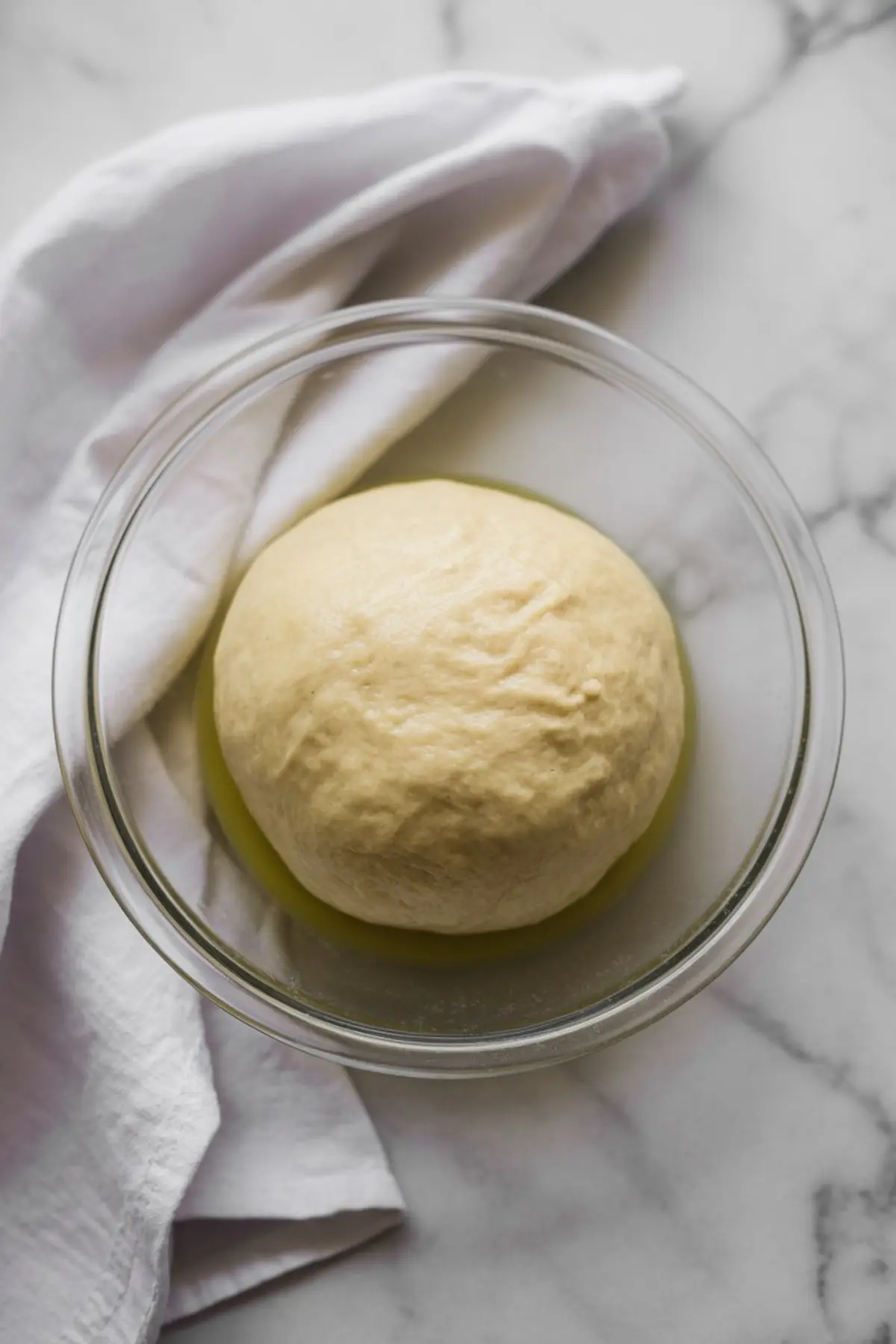 Risen dough ball in a glass bowl, resting on a marble surface and partially covered with a soft white cloth, ready for shaping.
