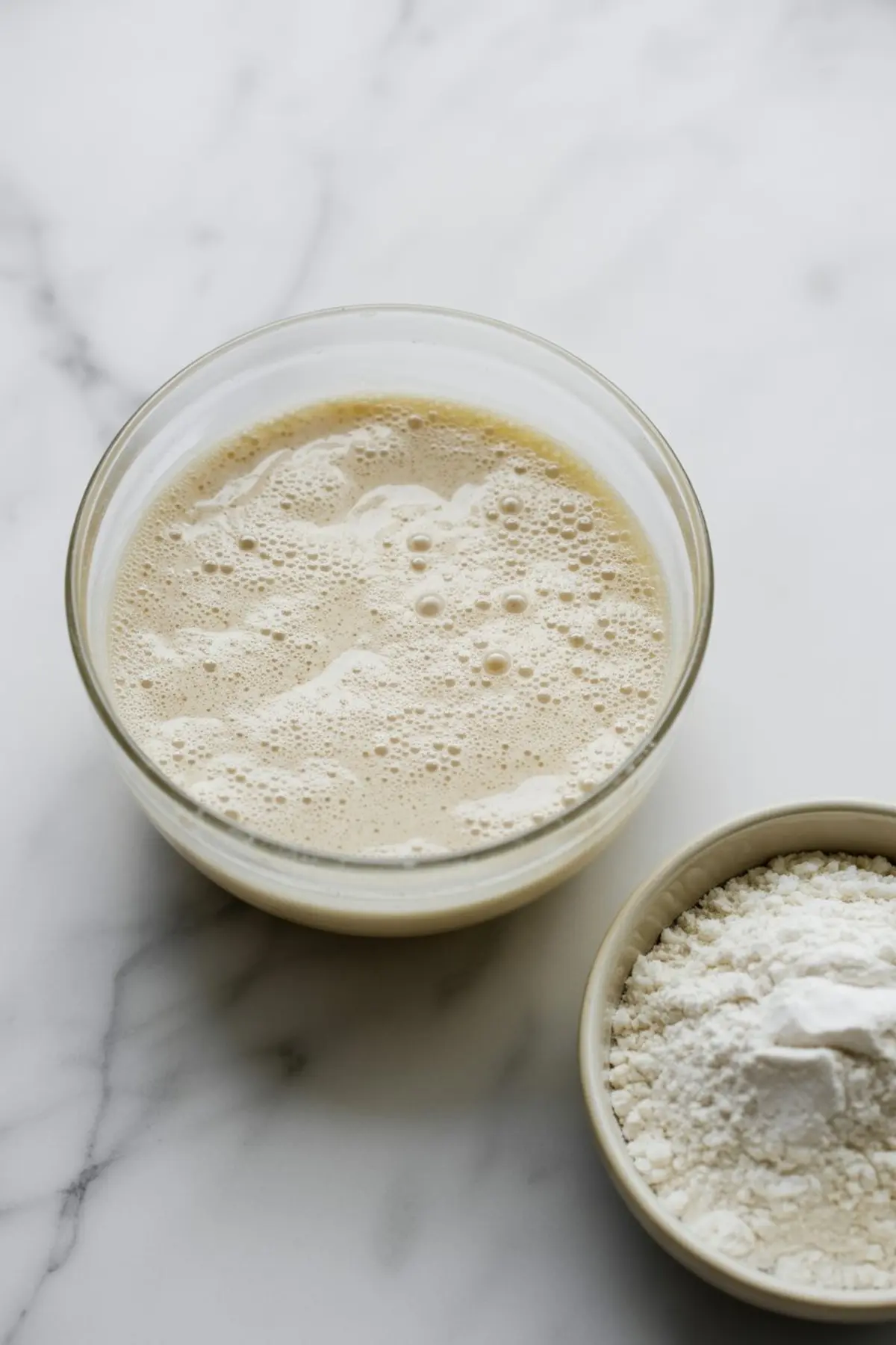 Close-up of bubbly active yeast mixture in a glass bowl next to a bowl of flour and baking powder, captured on a white marble background.
