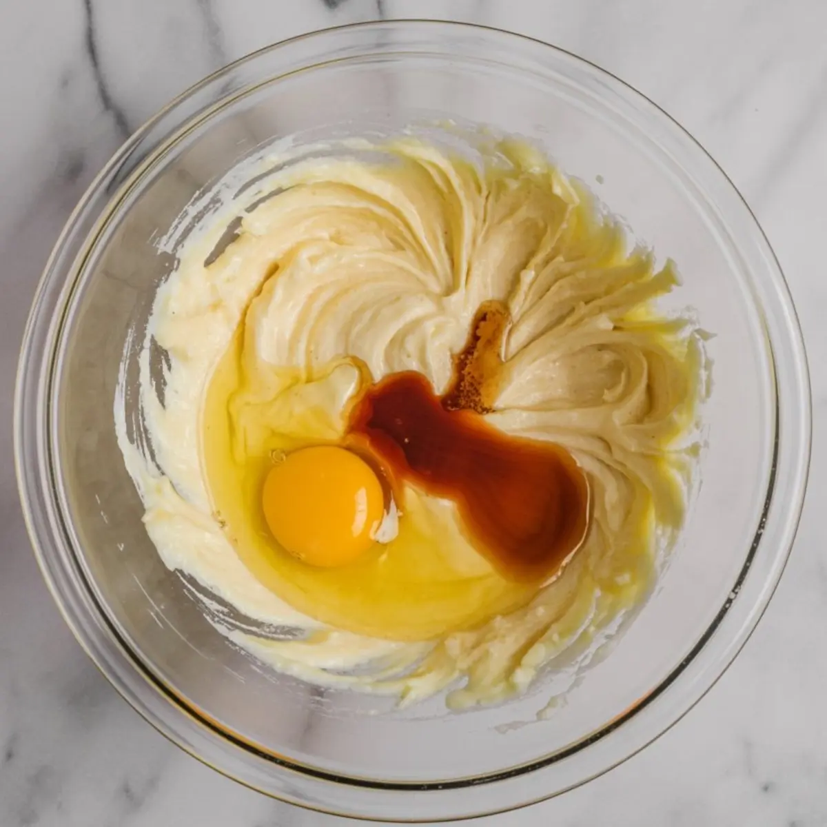 Glass mixing bowl with creamed butter and sugar, raw egg, and vanilla extract ready to be blended into sugar cookie dough.
