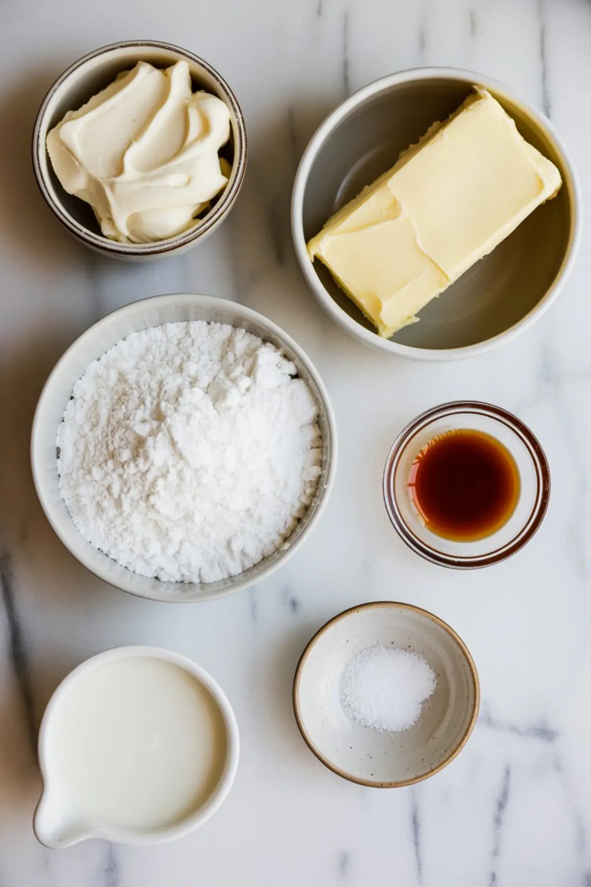 Flat lay of icing ingredients on marble: cream cheese, unsalted butter, powdered sugar, vanilla extract, milk, and a pinch of salt in ceramic bowls.
