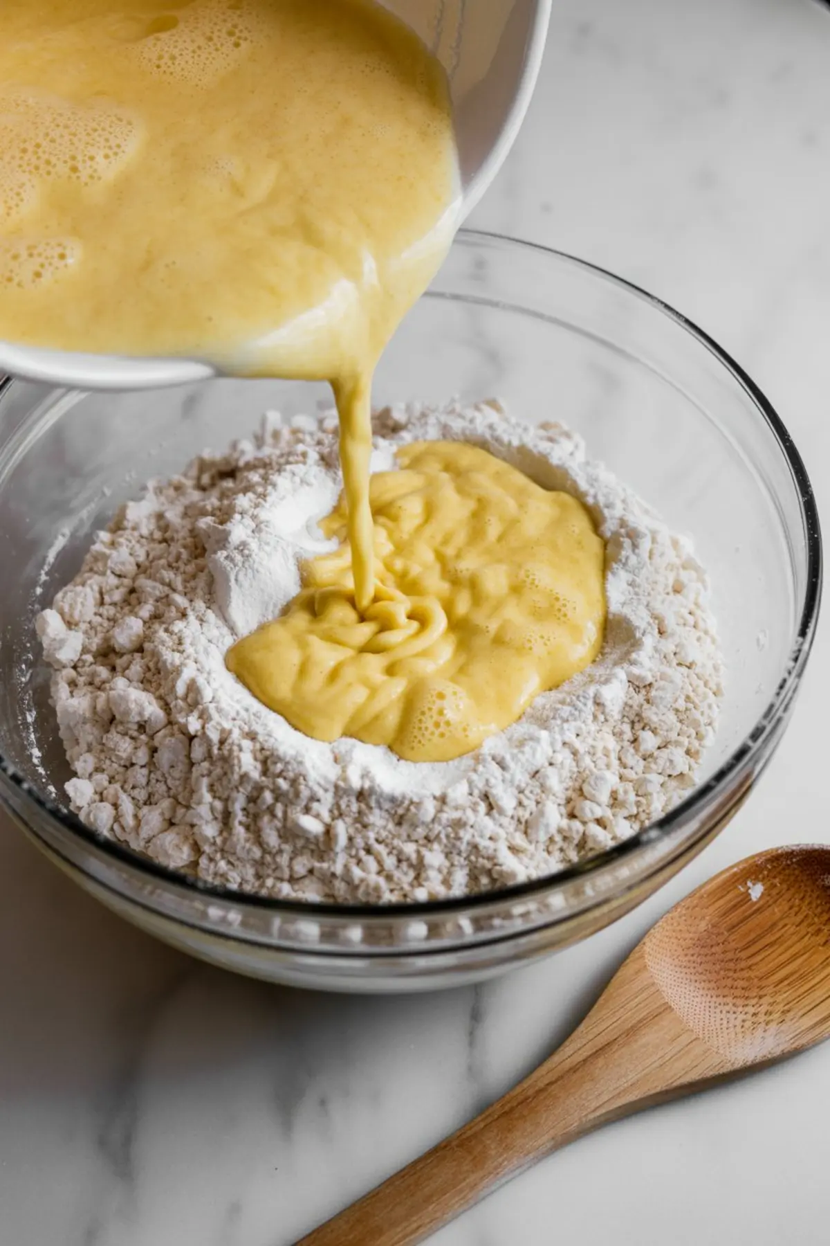 Smooth yellow egg mixture being poured into a bowl of dry ingredients including flour and sugar, showing the process of making muffin batter from scratch.
