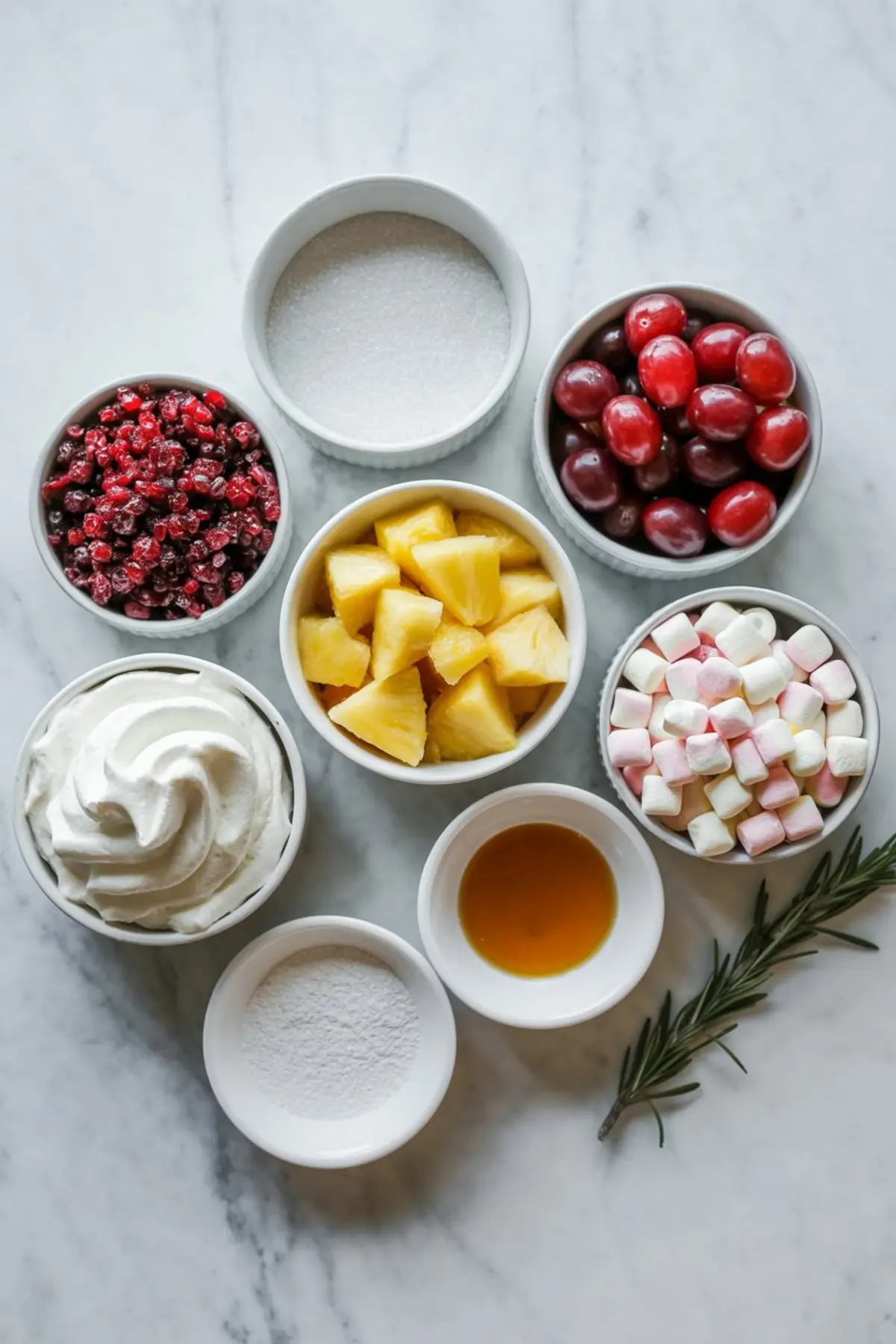 Flat lay of cranberry fluff salad ingredients in white bowls on marble background, including frozen cranberries, red grapes, pineapple chunks, mini marshmallows, whipped topping, granulated sugar, powdered sugar, vanilla extract, and a rosemary sprig.

