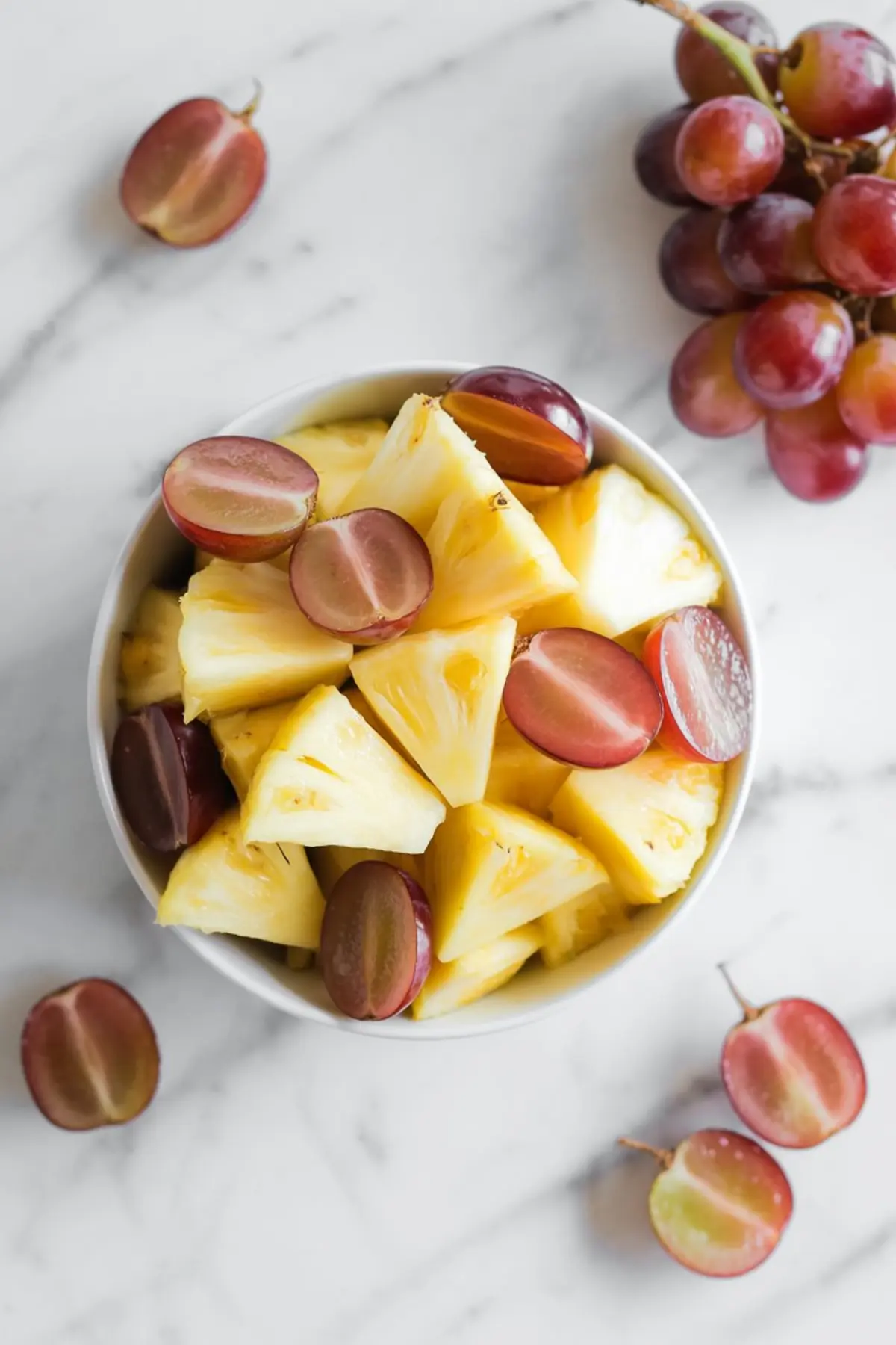 White bowl filled with fresh pineapple chunks and halved red grapes on a marble surface, showcasing vibrant tropical fruit mix for salads and sweet dishes.

