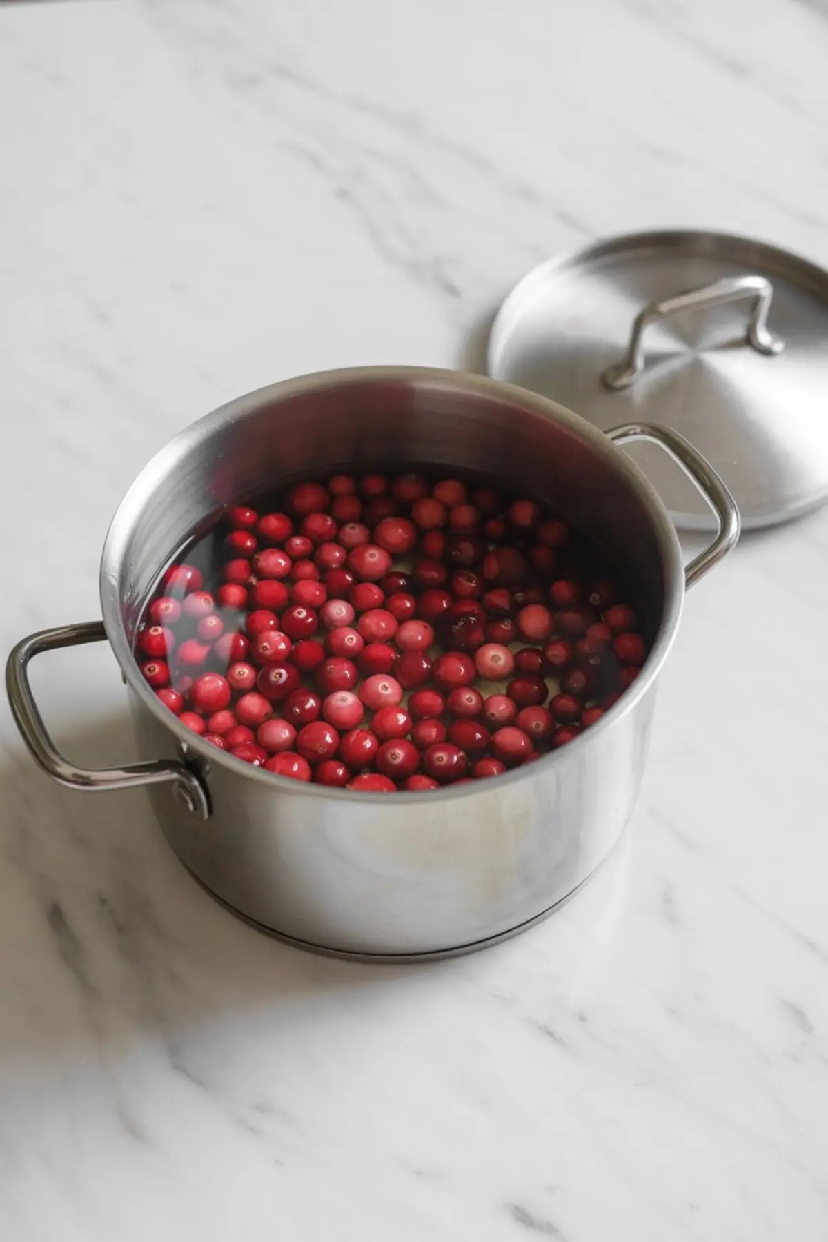 Large stainless steel pot filled with whole cranberries submerged in water, sitting on a marble countertop with the lid placed to the side, showing the beginning steps of homemade cranberry juice preparation.