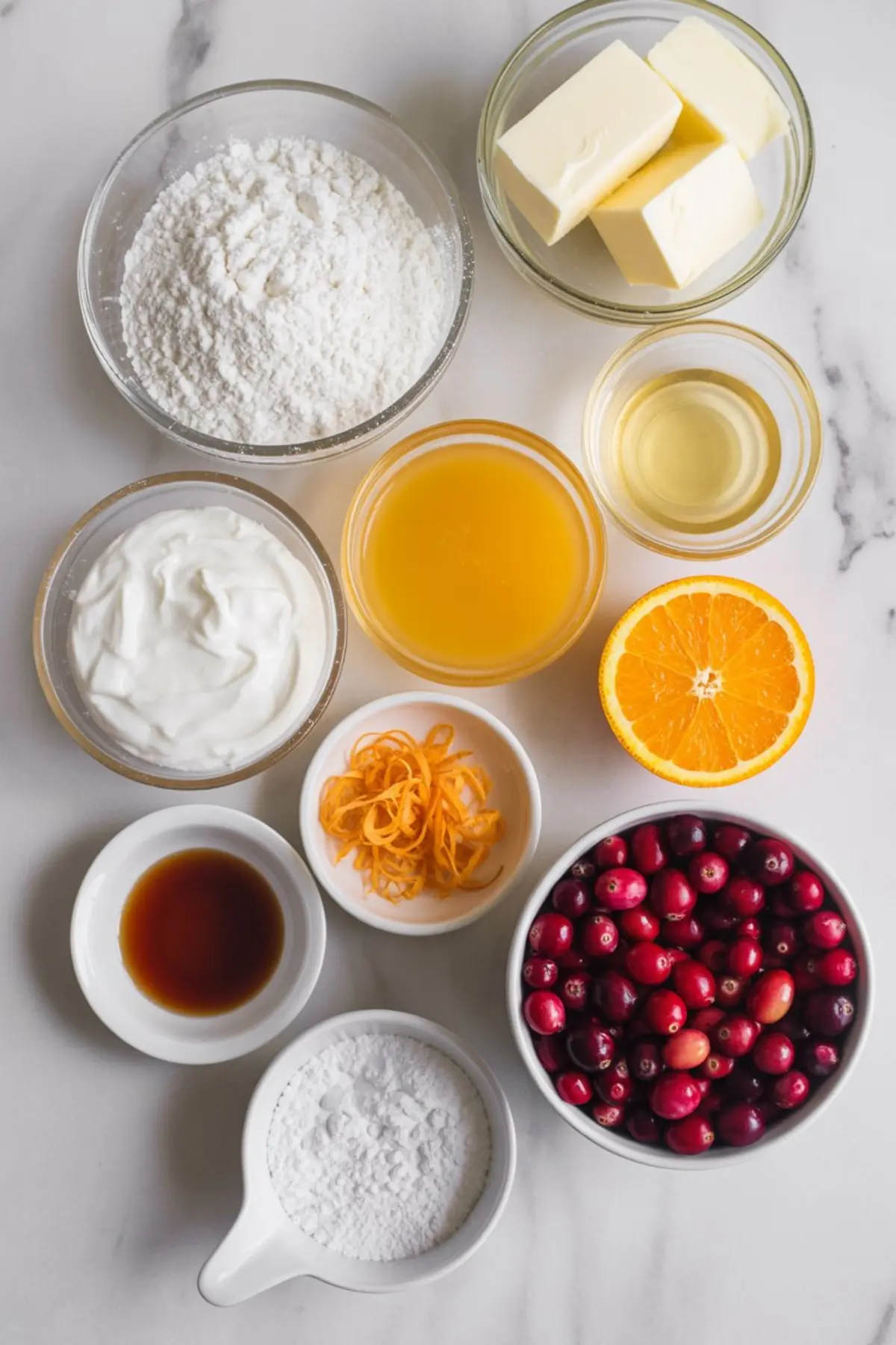 Overhead view of cranberry orange bread ingredients, including flour, butter, sour cream, orange juice, fresh cranberries, orange zest, vanilla extract, and powdered sugar, arranged on a marble counter.