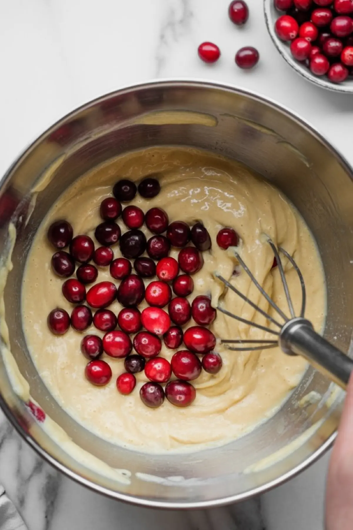 Stainless steel mixing bowl filled with cake batter and fresh cranberries being folded in with a whisk on a marble countertop.