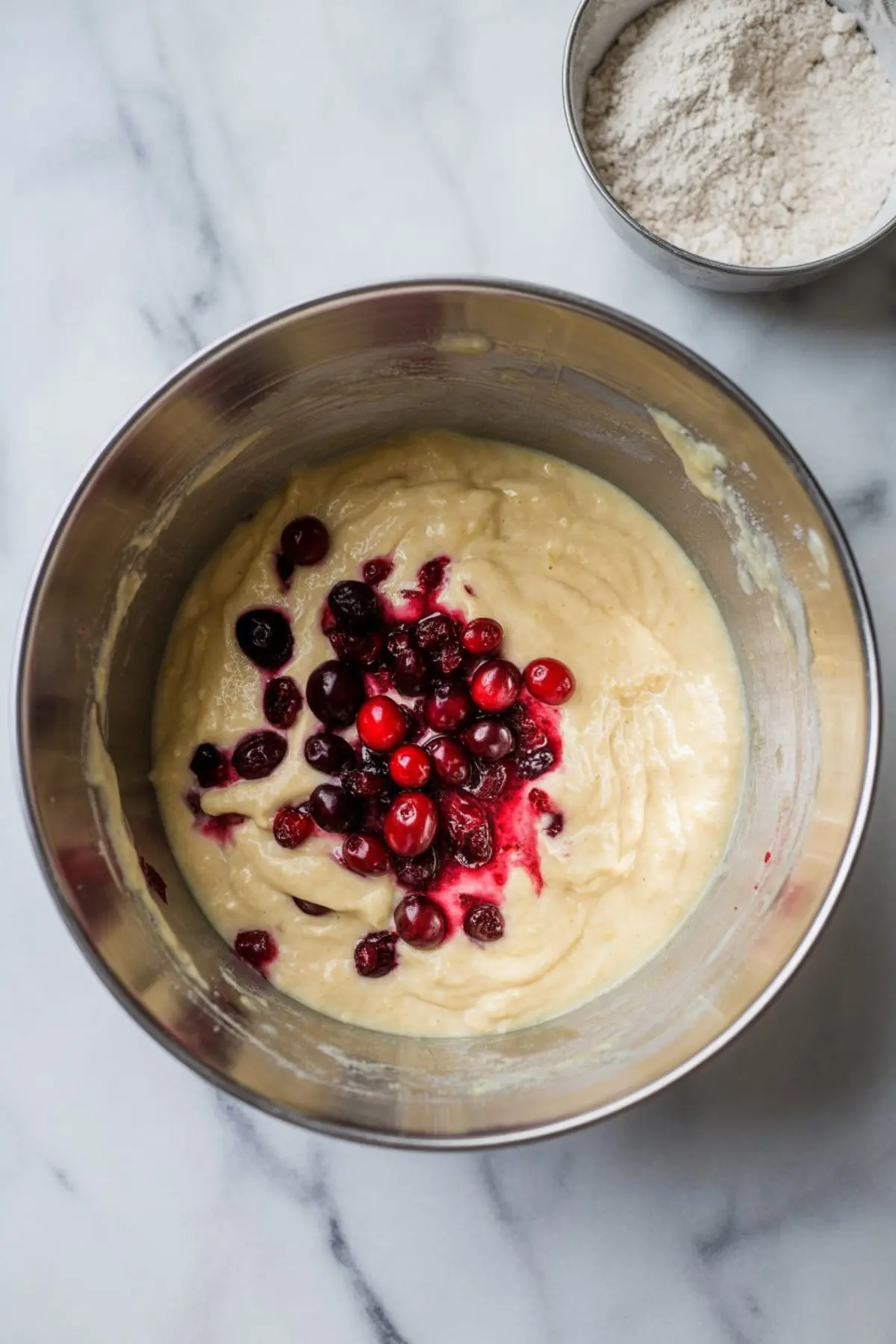 Mixing bowl filled with creamy muffin batter topped with fresh cranberries before folding them in.