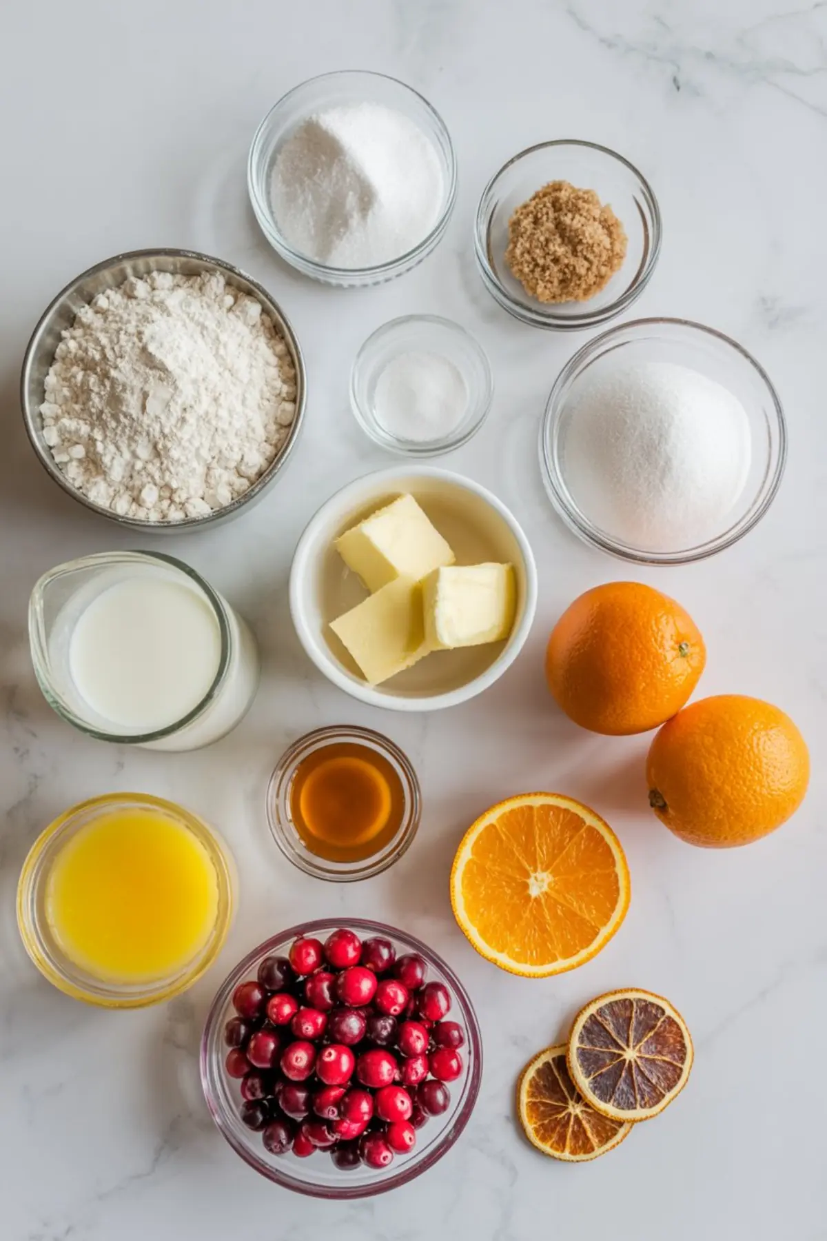 Overhead shot of baking ingredients for cranberry orange muffins, including flour, sugar, butter, milk, orange juice, vanilla, fresh cranberries, and whole oranges arranged on a marble surface.