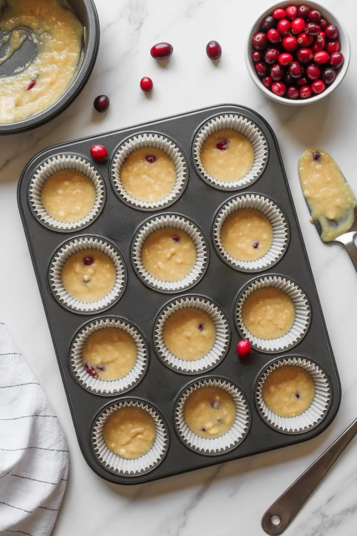 Muffin tin lined with paper cups filled with cranberry muffin batter, with a small bowl of fresh cranberries and a spatula on the side.