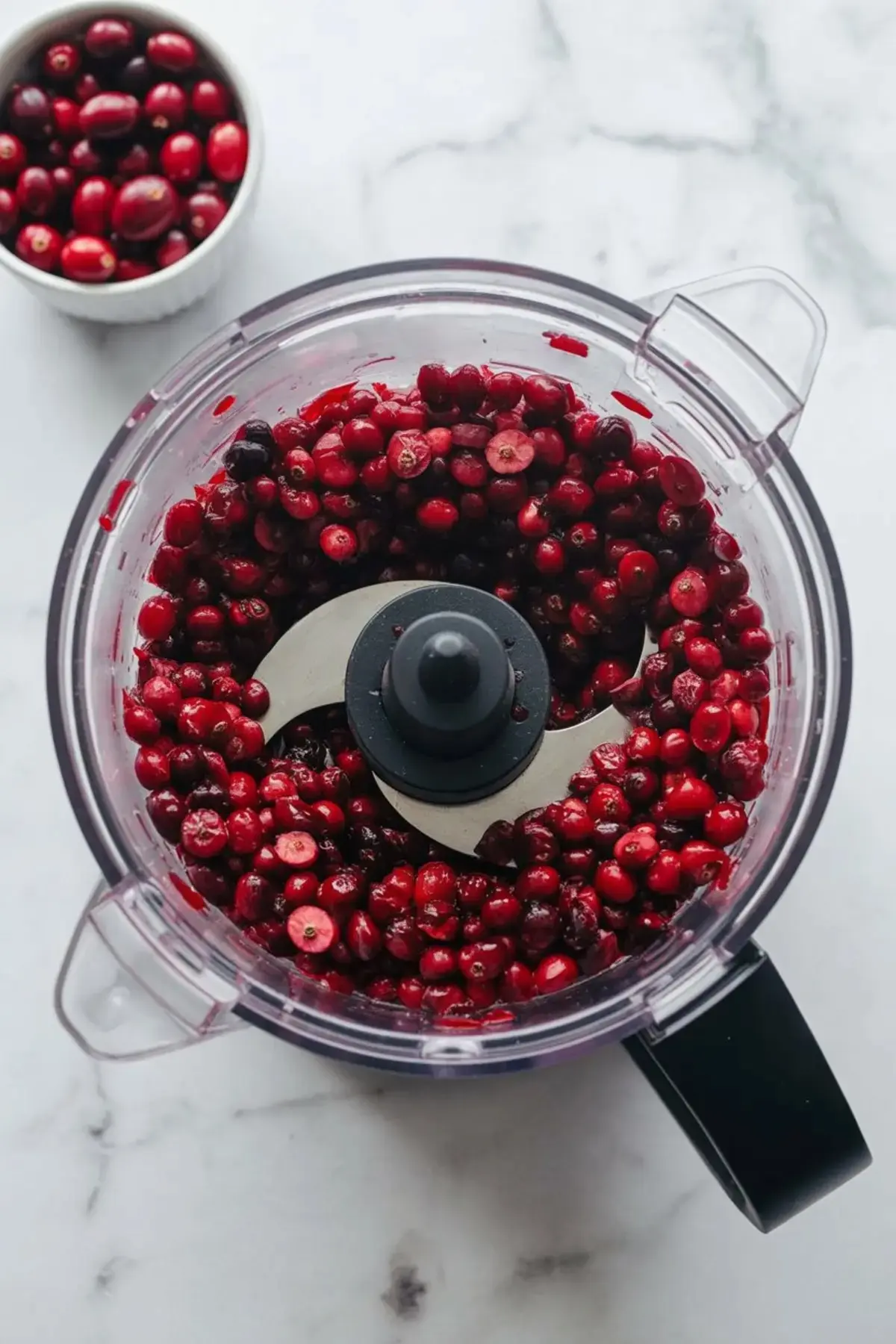 Fresh cranberries inside a food processor with a sharp blade, ready to be chopped on a white marble countertop, with an extra bowl of cranberries nearby.