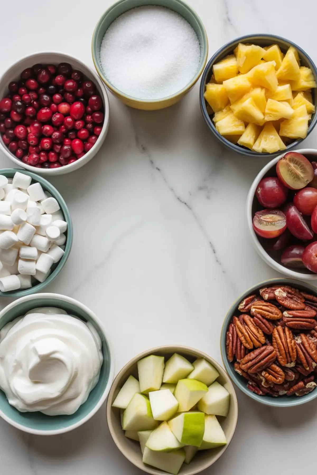 Overhead view of bowls with ingredients for cranberry salad, including fresh cranberries, sugar, pineapple chunks, grapes, pecans, green apple cubes, whipped cream, and mini marshmallows on a white marble surface.