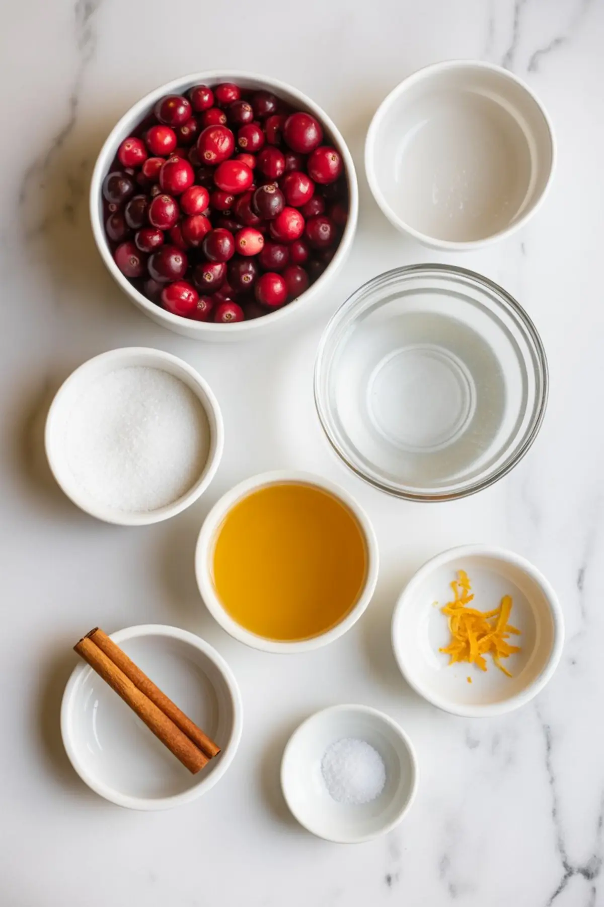 Flat lay of cranberry sauce ingredients in white bowls, featuring fresh cranberries, granulated sugar, orange juice, water, cinnamon sticks, salt, and orange zest on a marble background.

