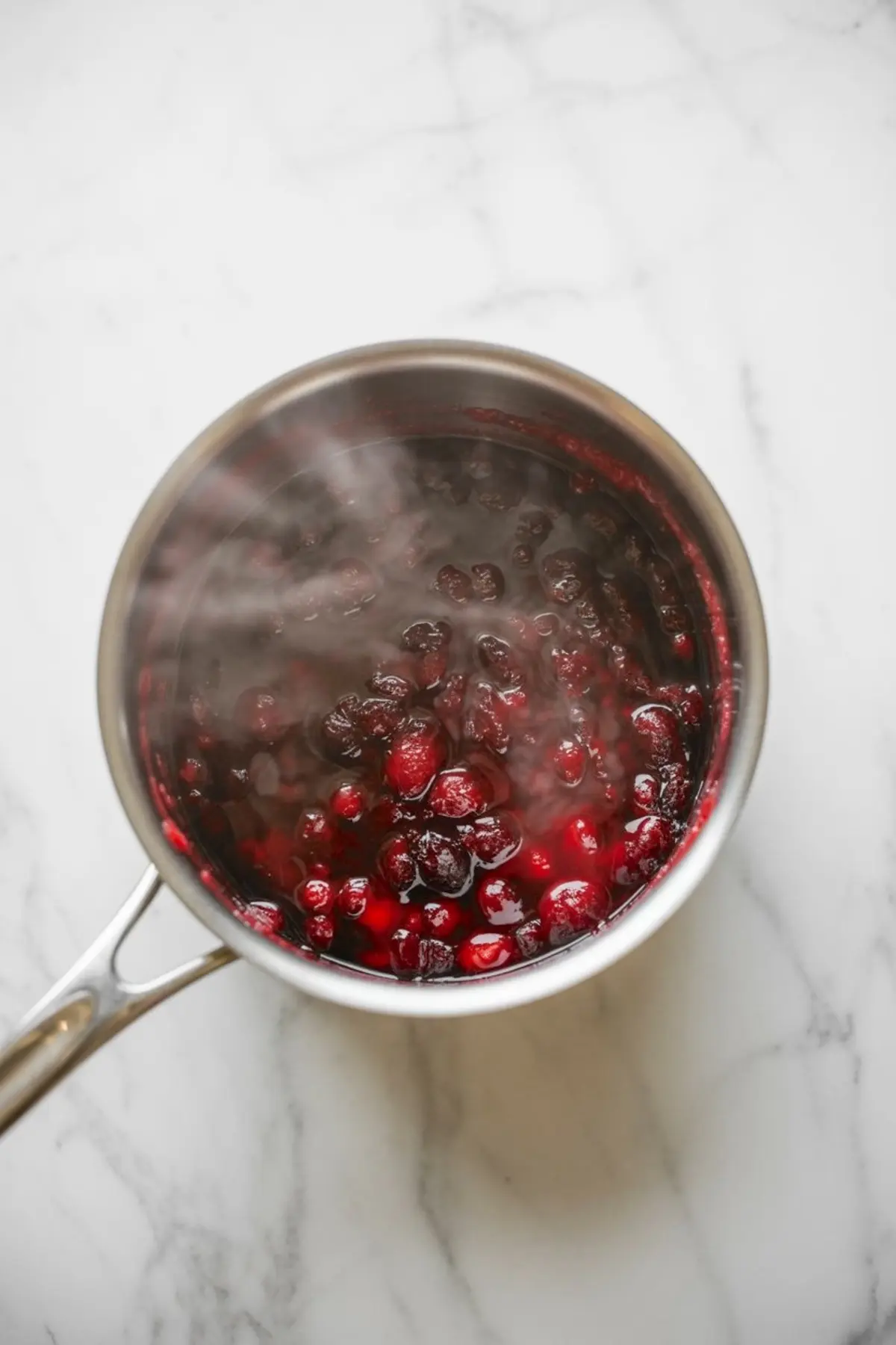 Saucepan filled with simmering cranberry sauce, with whole cranberries bursting in a thick red liquid on a white marble surface.
