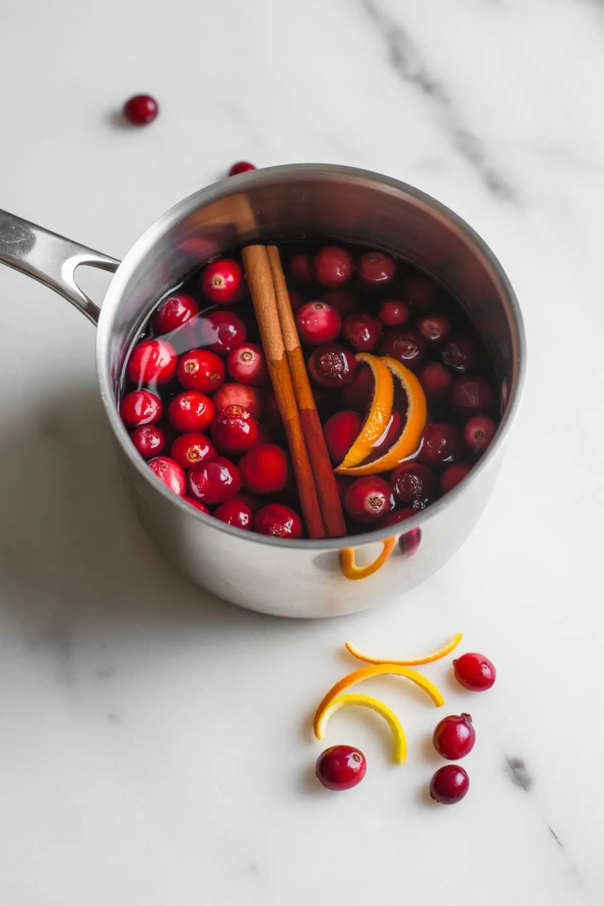 Uncooked cranberry sauce mixture in a stainless steel pot, with floating fresh cranberries, cinnamon sticks, and orange slices on a marble countertop.
