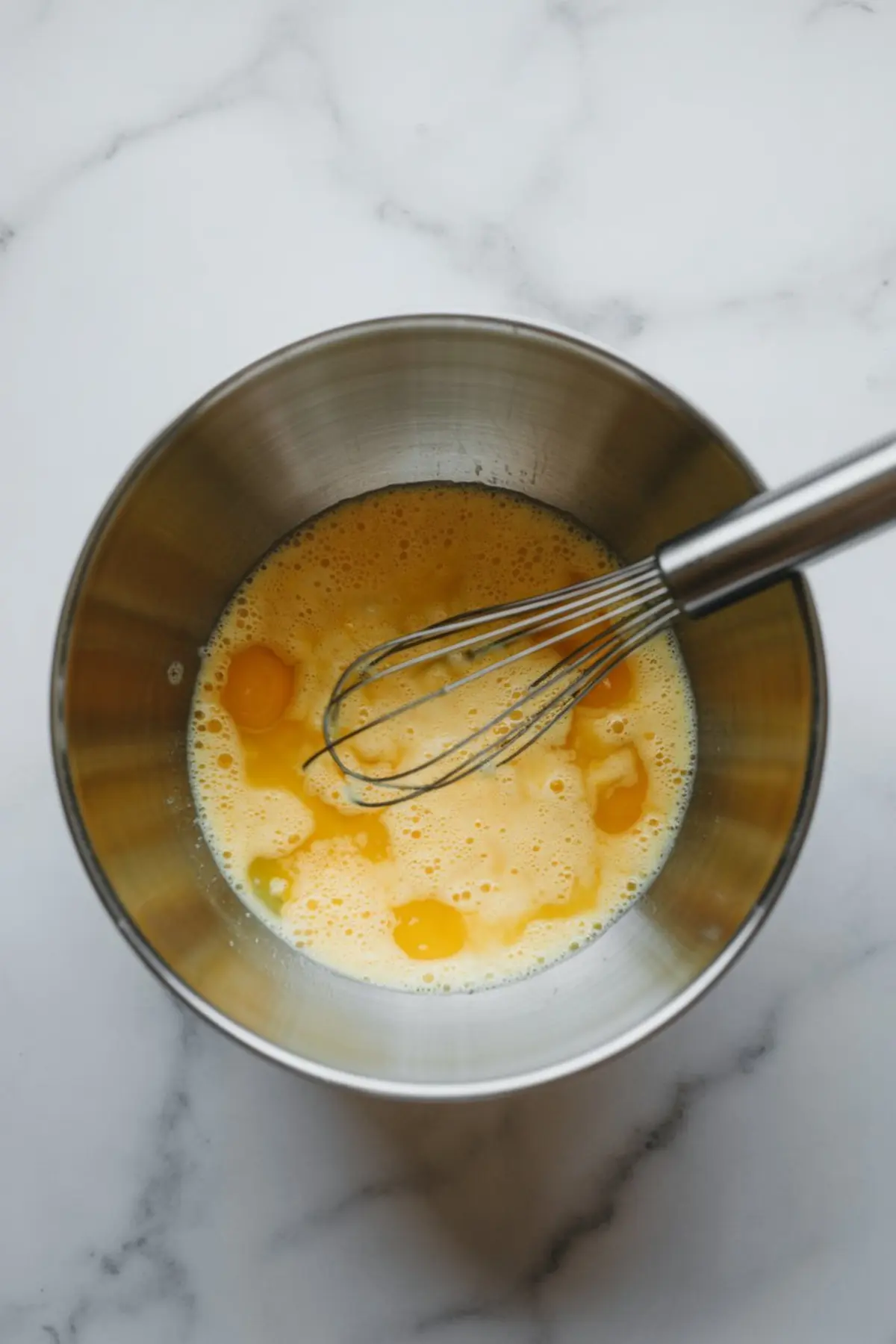 Whisk resting in a metal bowl with lightly beaten eggs and milk, ready for blending into a silky custard filling.
