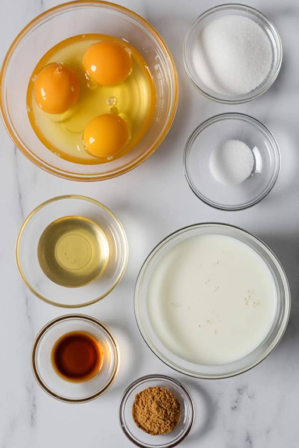 Flat lay of custard pie ingredients in clear glass bowls on a marble background, featuring eggs, sugar, milk, vanilla extract, ground nutmeg, and oil.

