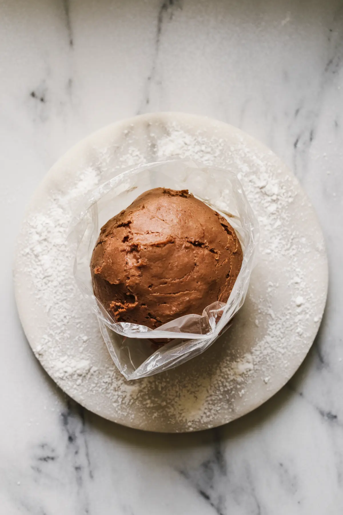 Ball of chocolate tart dough wrapped in plastic wrap resting on a floured round board, prepared for chilling before rolling.
