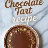 Baked chocolate tart with glossy ganache, garnished with gold flakes, crushed pistachios, and rose petals, labeled “Dubai Chocolate Tart recipe.”