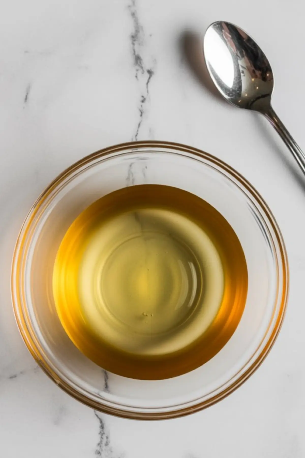 Close-up of a glass bowl filled with light corn syrup next to a silver spoon on a white marble surface, an essential ingredient in making edible fake blood.