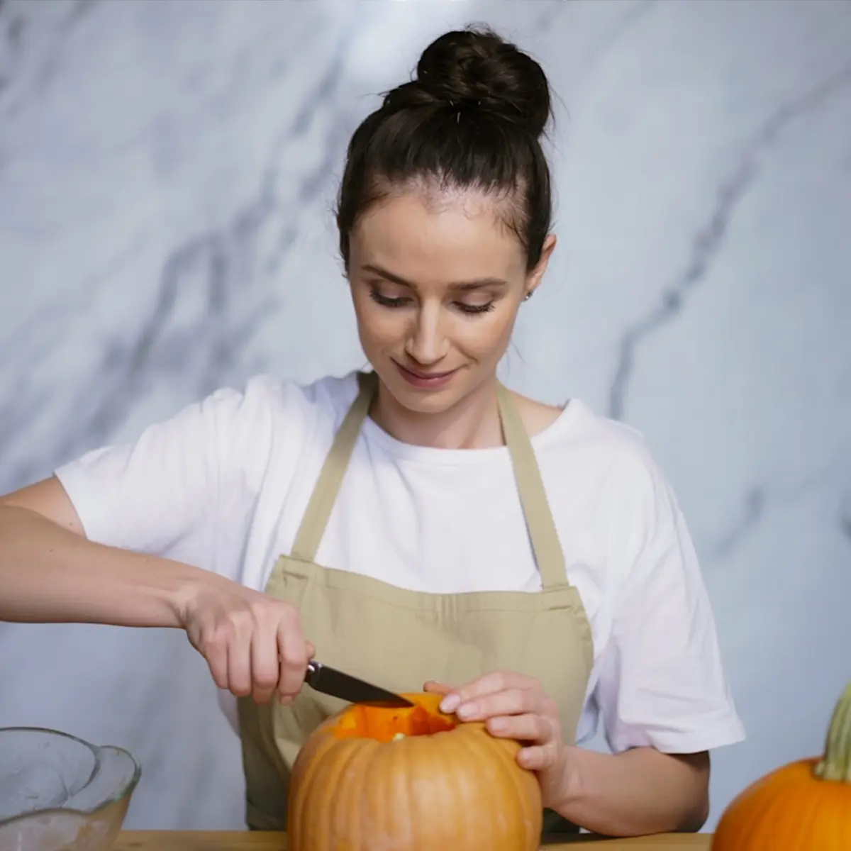 Emma in a beige apron cutting open a fresh pumpkin on a wooden table against a marble background.